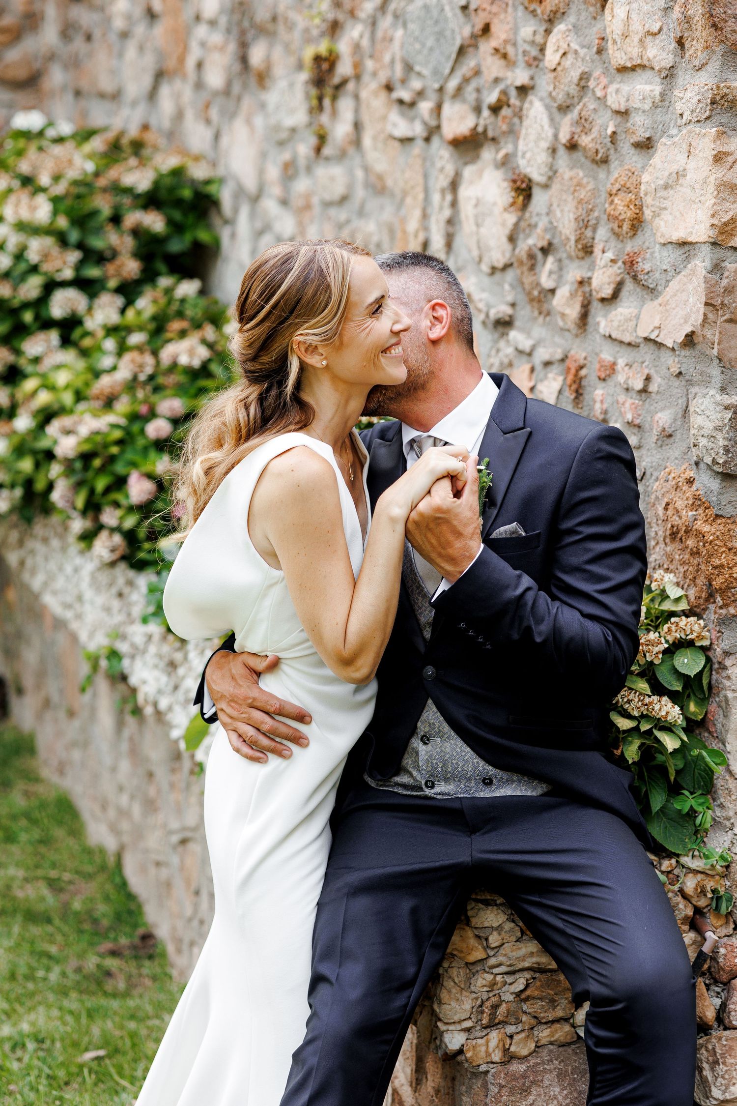 A bride and groom share a romantic kiss against a rustic stone wall adorned with blooming hydrangeas at their wedding.
