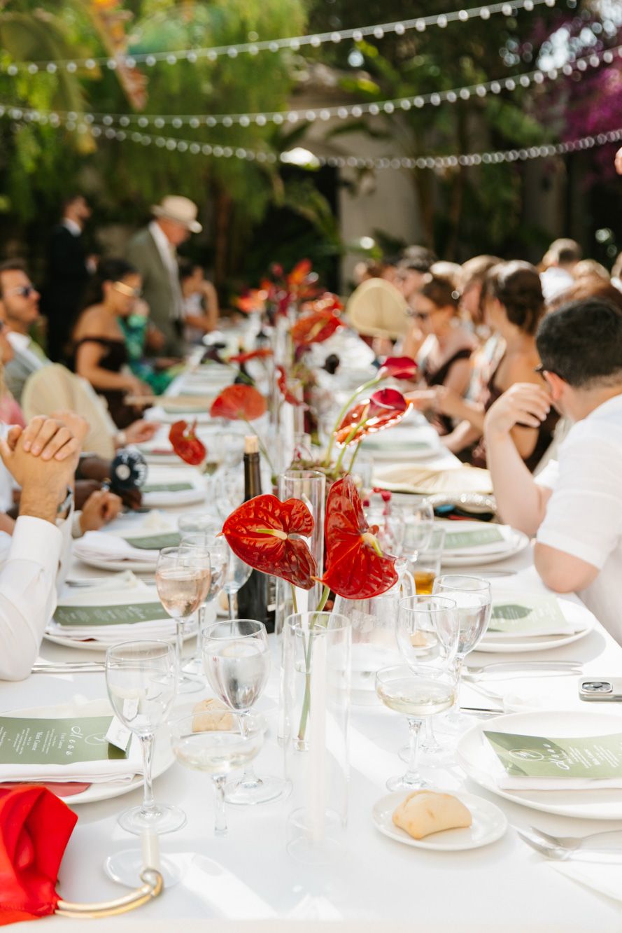 Elegant outdoor wedding reception table with red anthurium centerpieces, white linens, and guests dining under string lights.