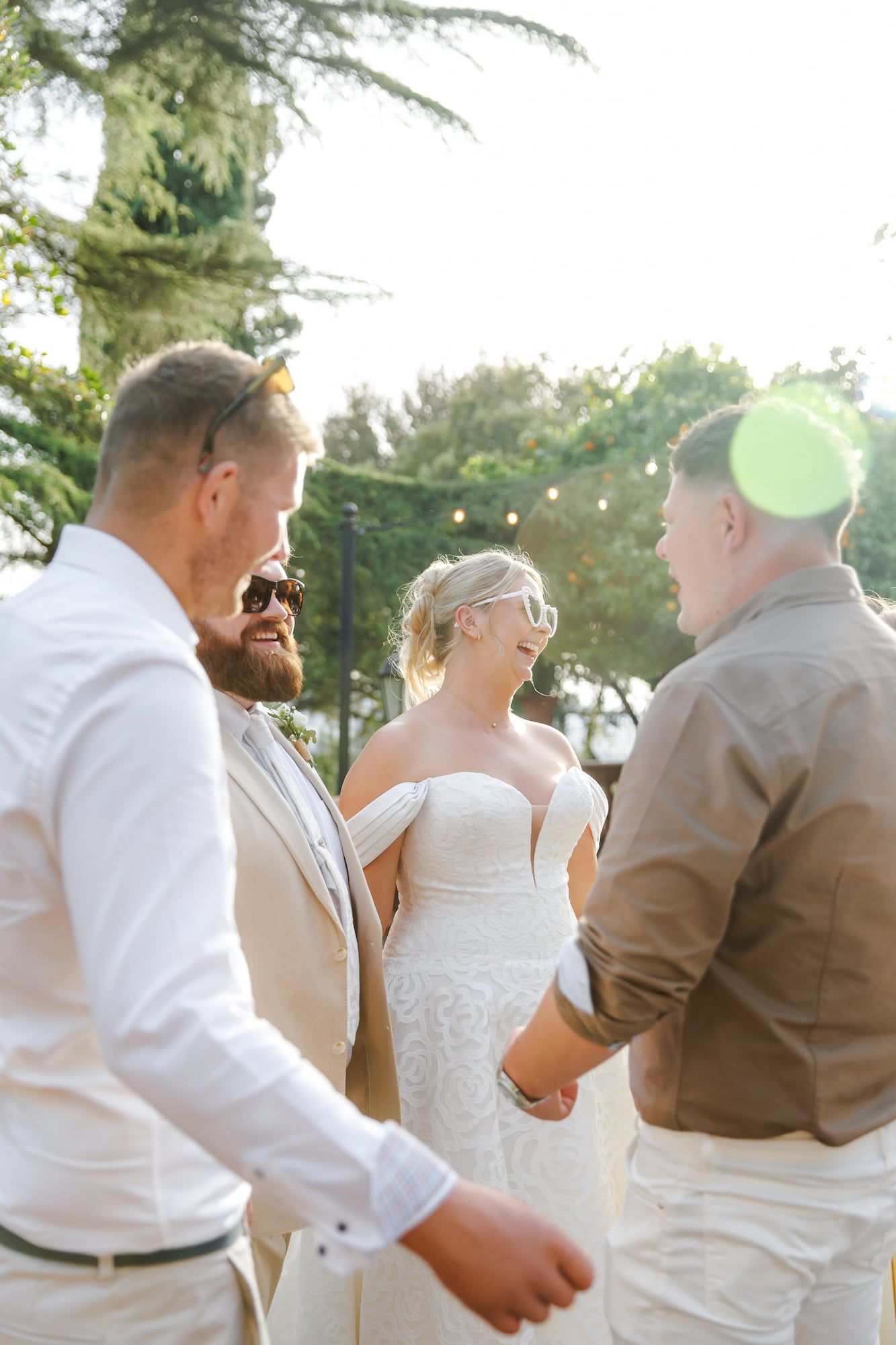 Bride in strapless lace gown laughs with groom in tan suit and guests at sunny outdoor wedding ceremony.