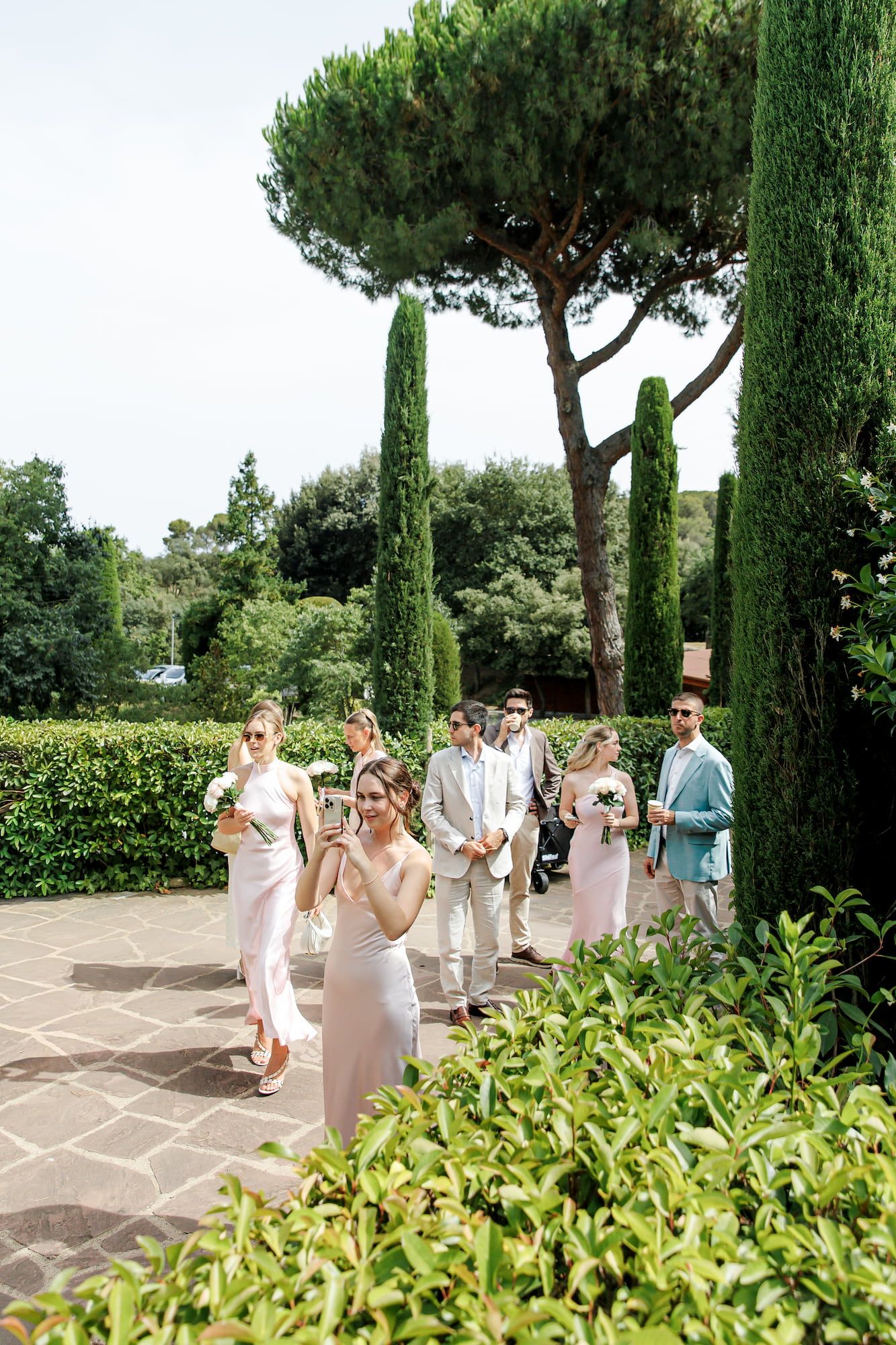 Elegantly dressed wedding guests stroll through a lush Italian garden with tall cypress trees on a sunny day.