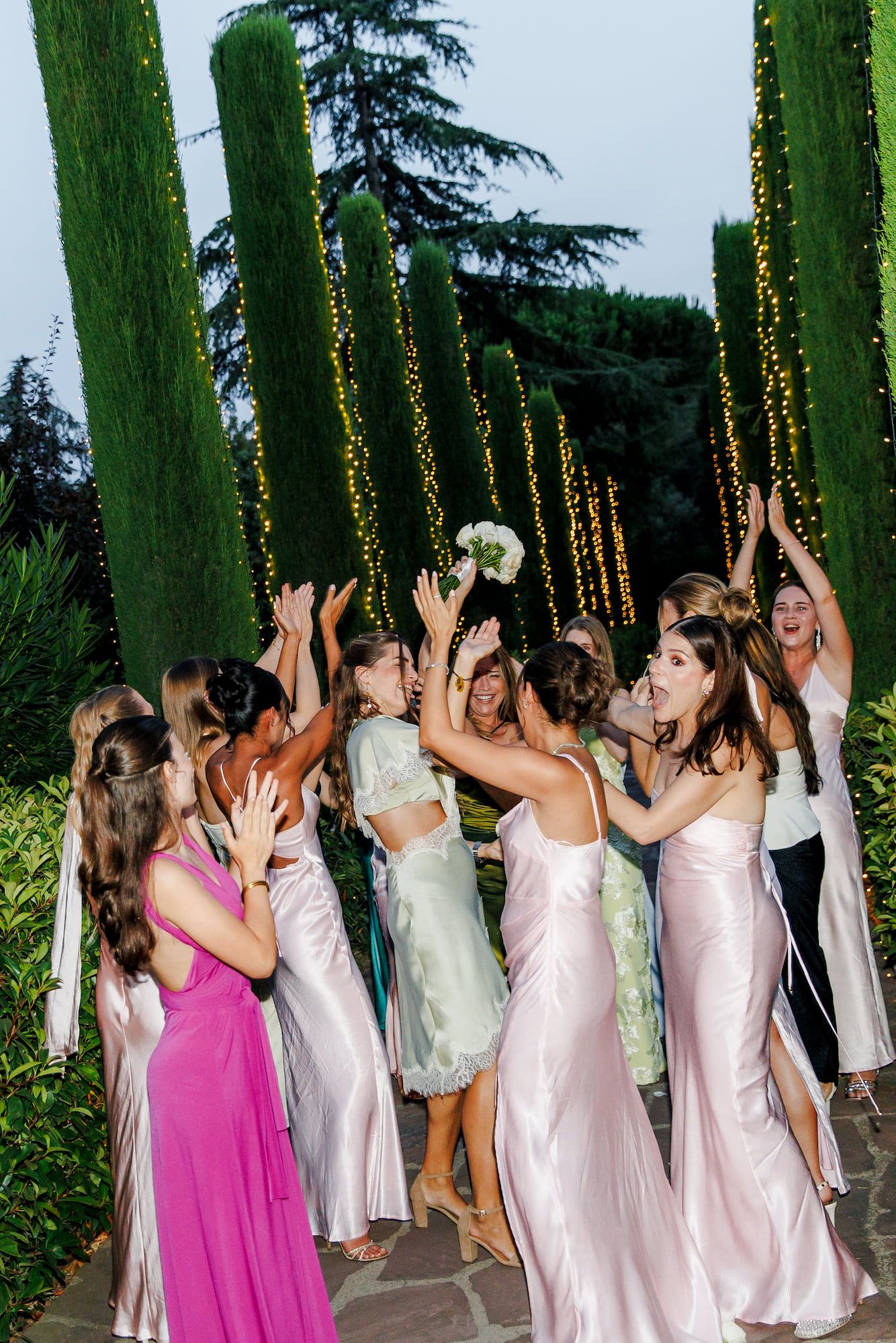 Bridesmaids in pink satin gowns reach for a bouquet toss at an outdoor wedding reception with lit cypress trees.