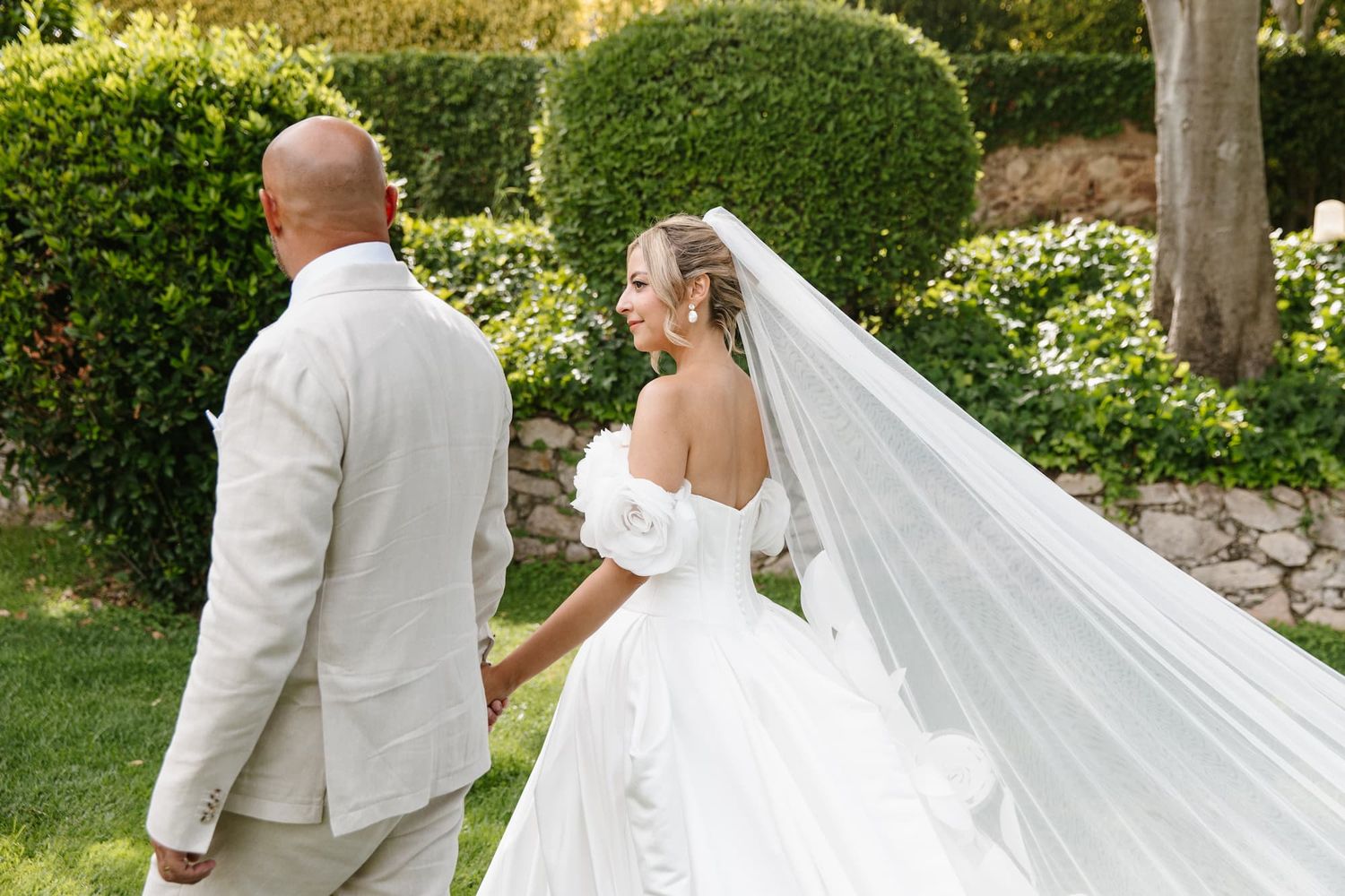 Bride in off-shoulder gown with veil holds hands with groom in beige suit outdoors in lush garden setting.