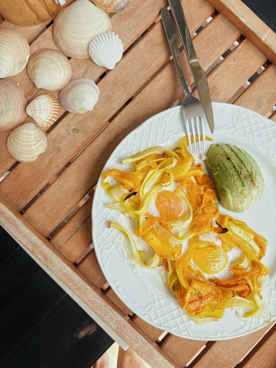 A white plate with yellow pasta and avocado on a wooden tray with seashells, fork and knife beside it.