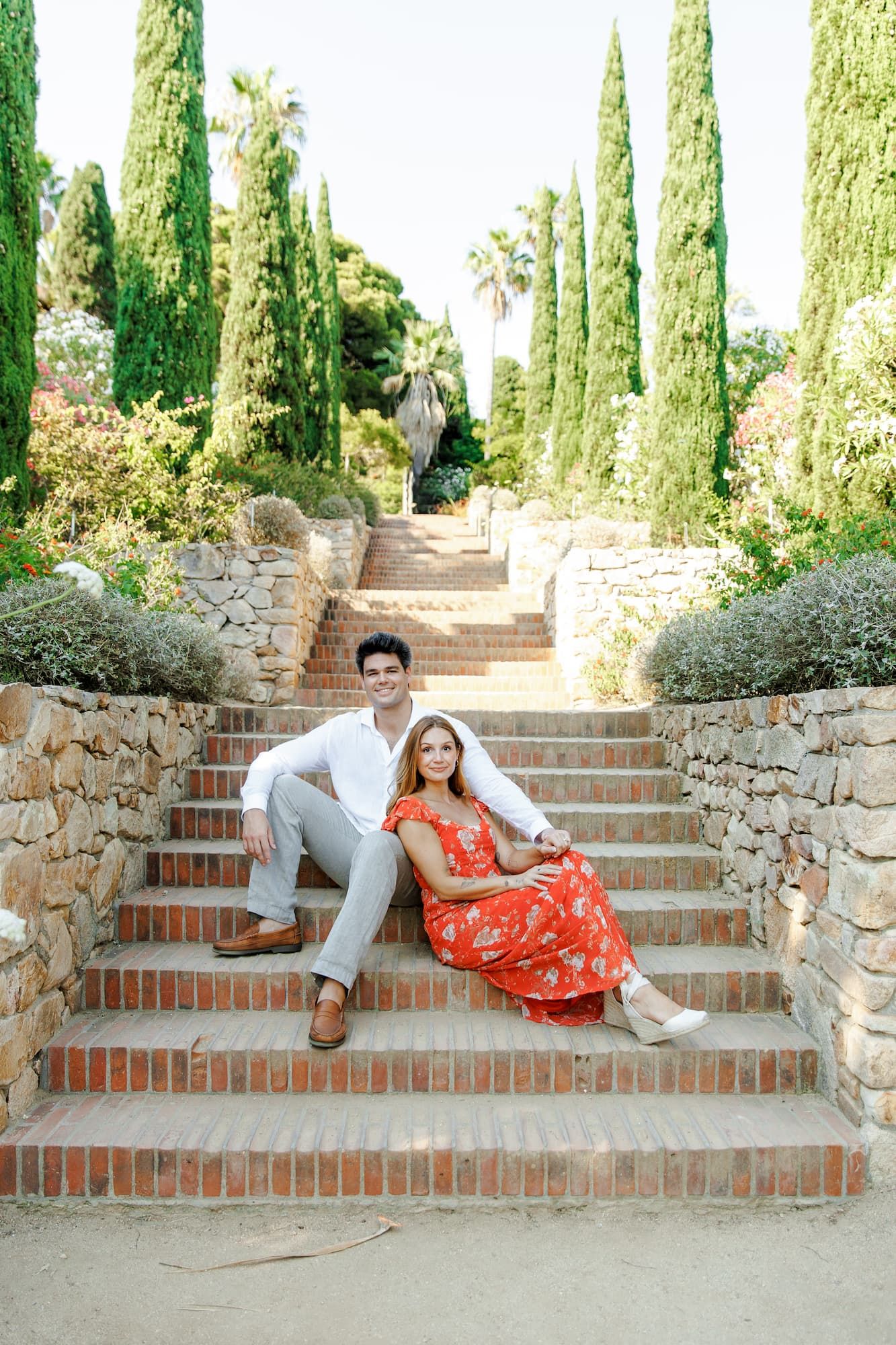 Couple sitting on brick garden steps surrounded by cypress trees, woman in red floral dress, man in white shirt.