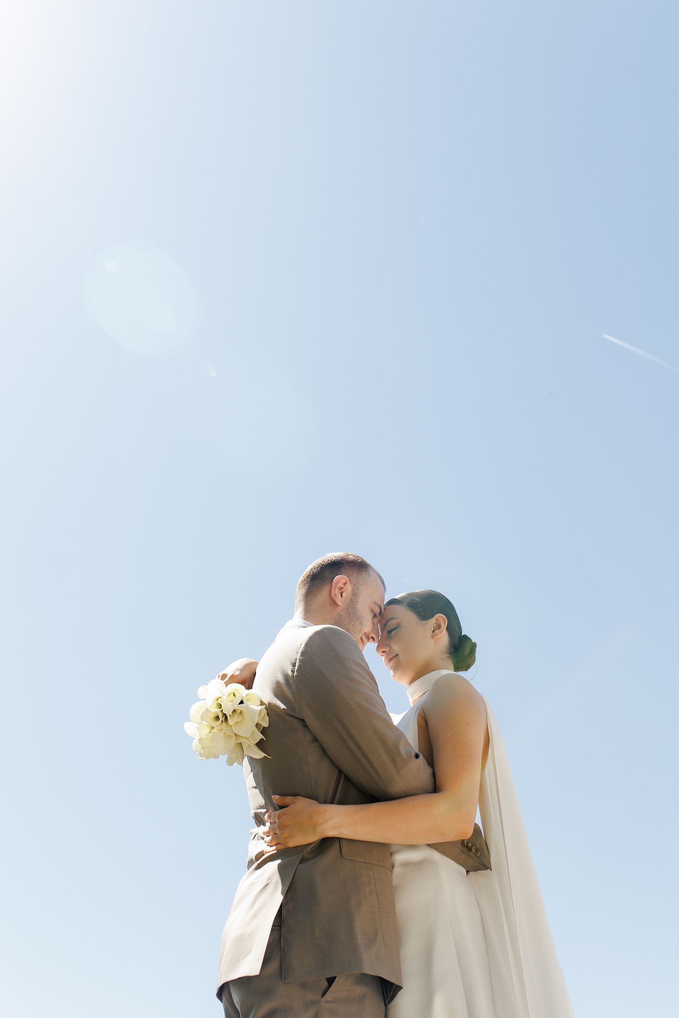 A romantic couple embraces during their outdoor wedding ceremony against a bright blue sky background.