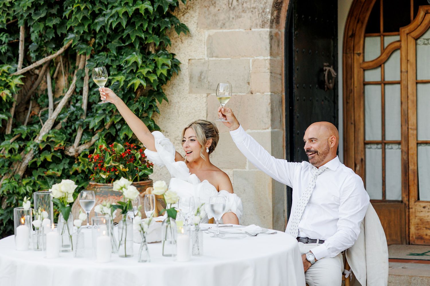 A bride and groom raise champagne glasses in a toast at their outdoor wedding reception table adorned with white floral arrangements.