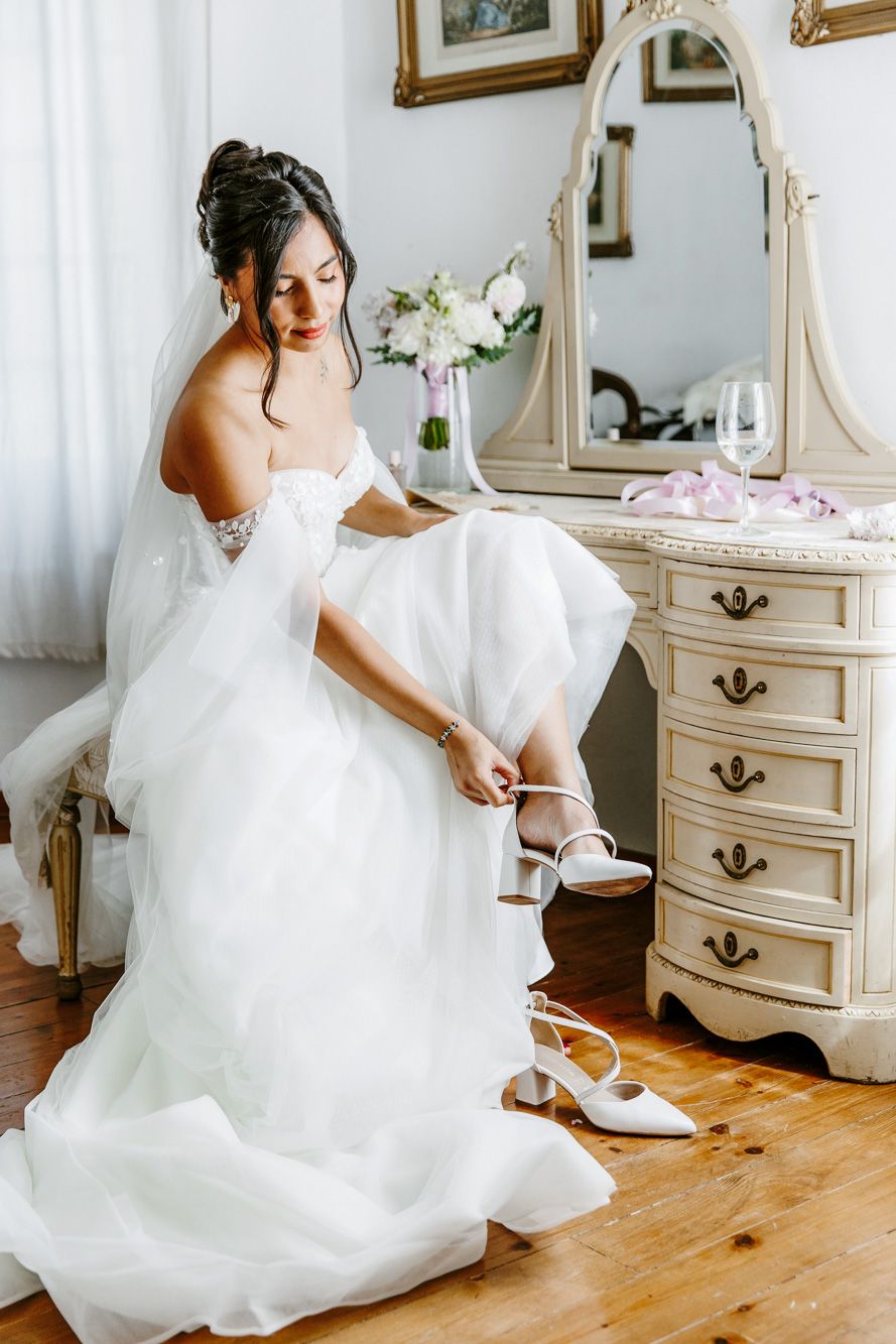 Bride in white gown adjusting her heels beside a vintage dresser with mirror on her wedding day.