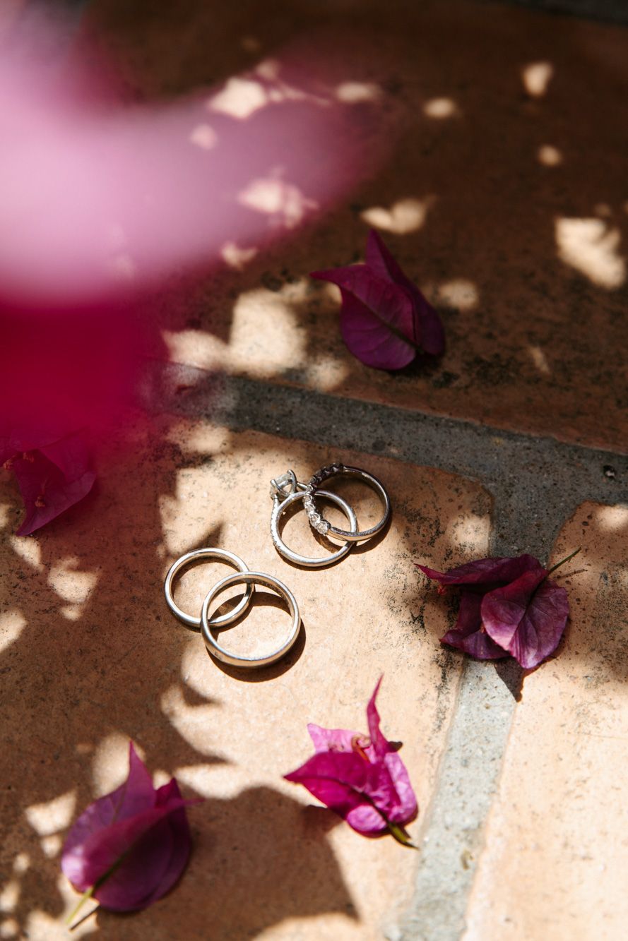 Three gold wedding rings and a diamond engagement ring arranged on terracotta tiles with purple bougainvillea petals in sunlight.