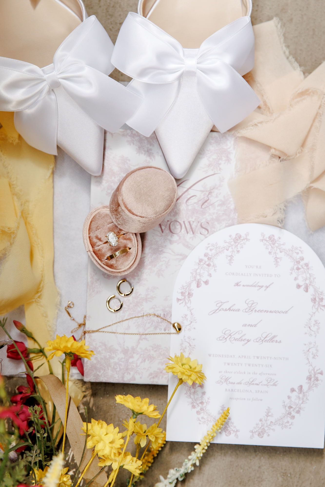 Elegant wedding flat lay with floral invitations, ring box, gold bands, white bow shoes, and yellow flowers on a neutral background.