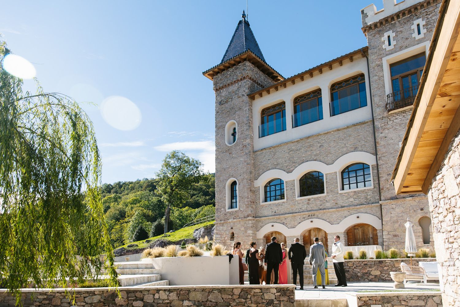 Wedding guests gather on a stone terrace outside a historic castle-style building with a pointed tower, surrounded by green hills and trees.