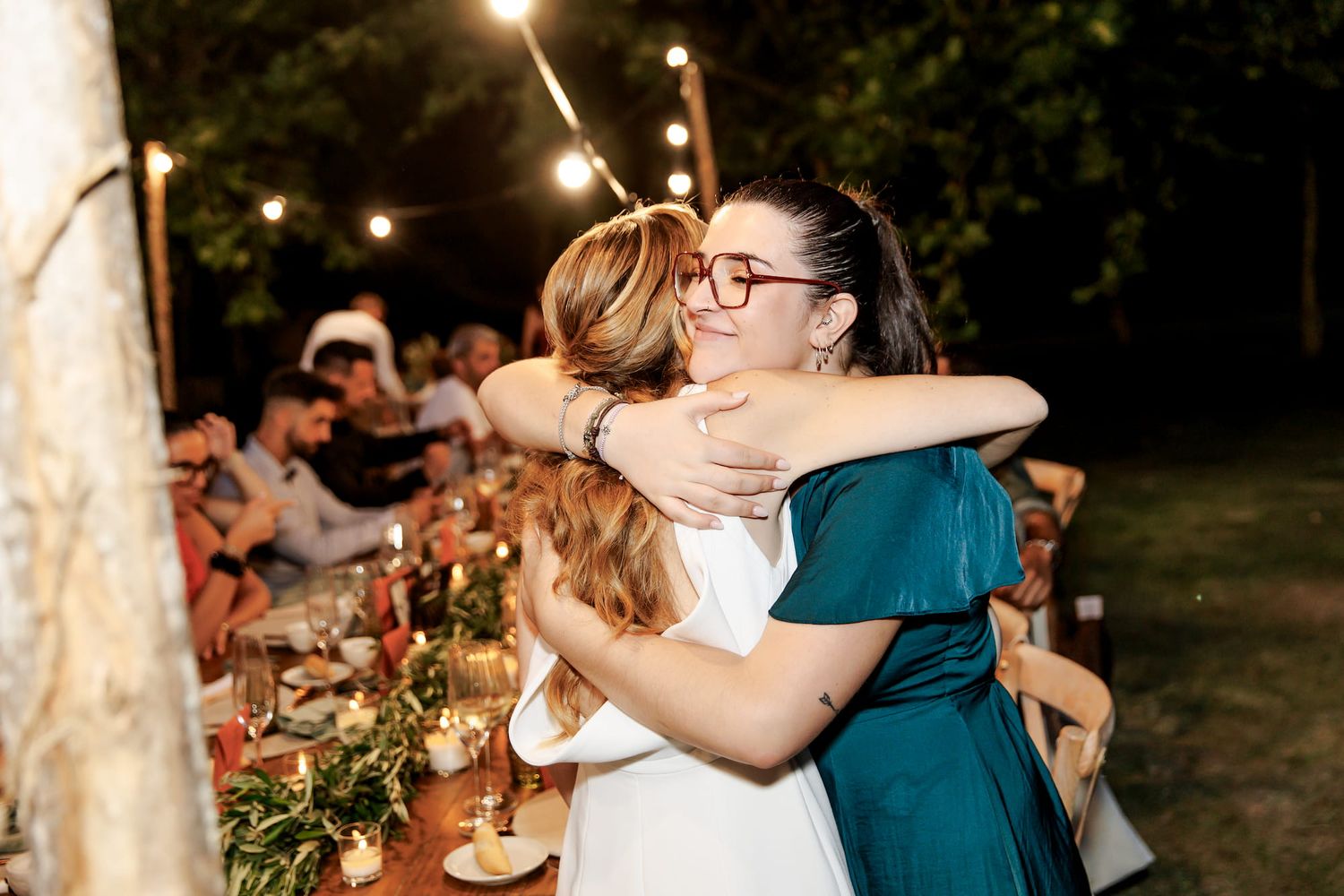 Two women share a warm hug at an outdoor wedding reception dinner under string lights at night.