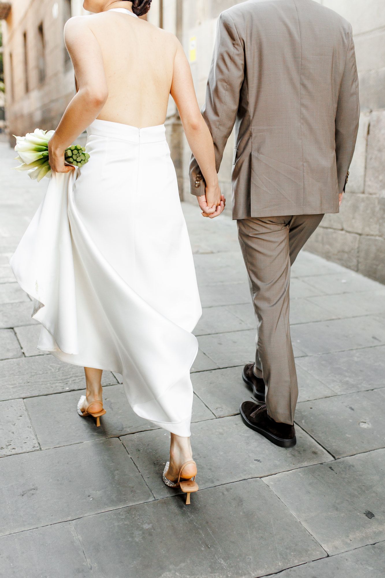 A newlywed couple walking together on the stone pavement in the Gothic Quarter in Barcelona, holding hands.