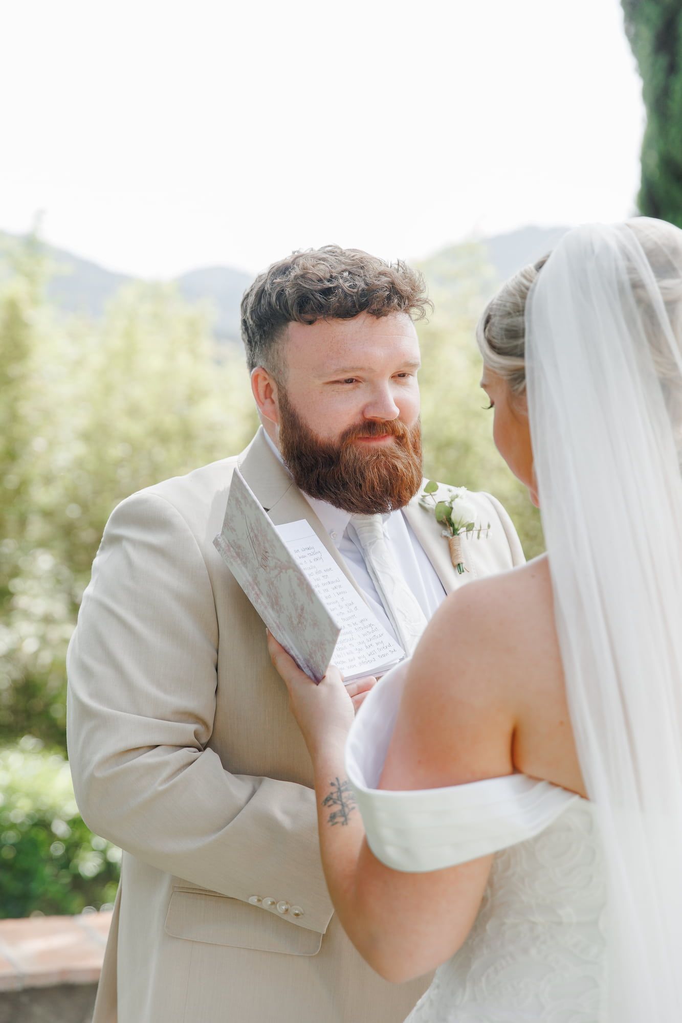 Bearded groom in tan suit reads vows to bride in white dress and veil during outdoor wedding ceremony.