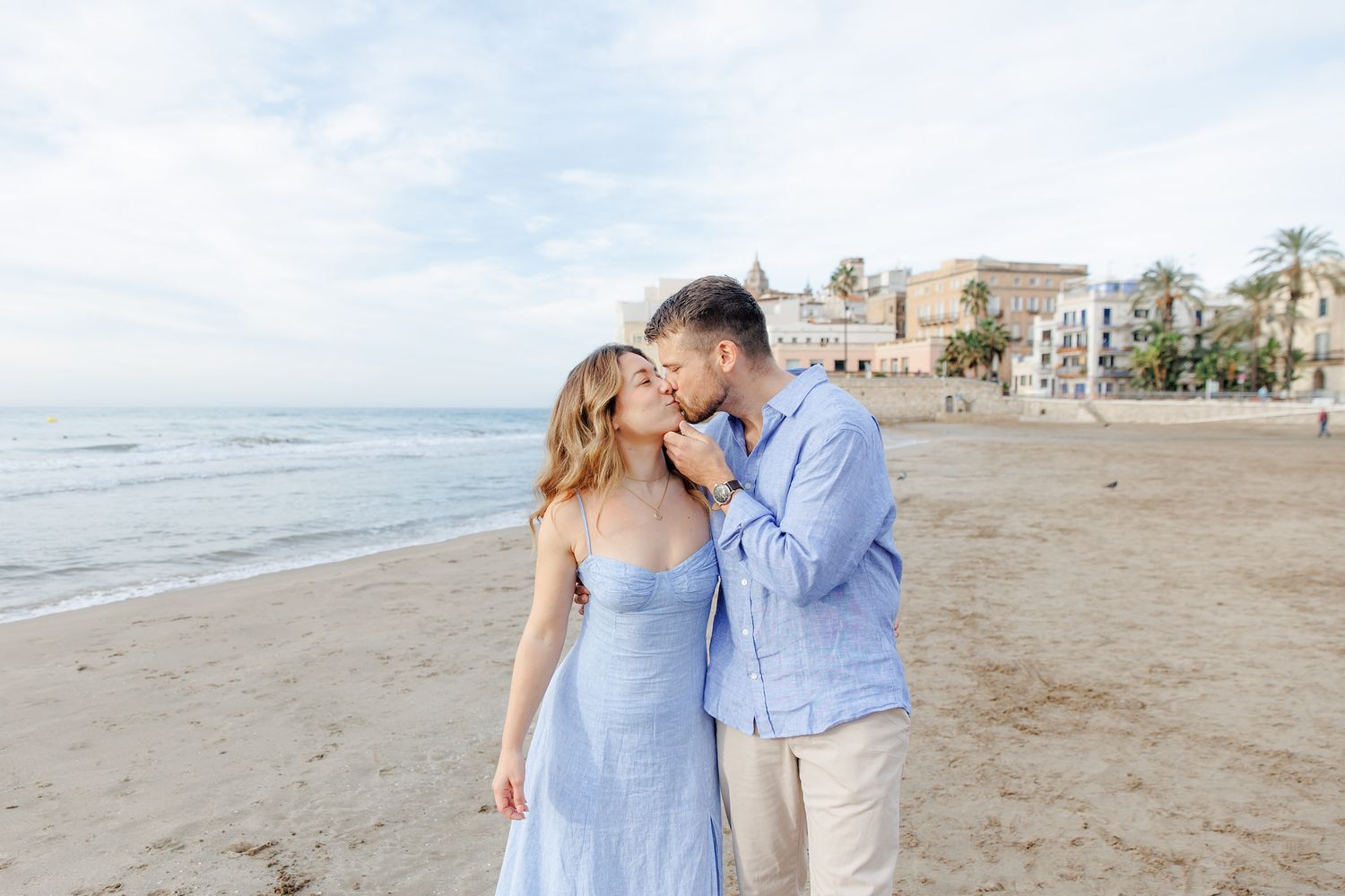 A couple in coordinating blue and white outfits share romantic moments together on a sandy beach at sunset.