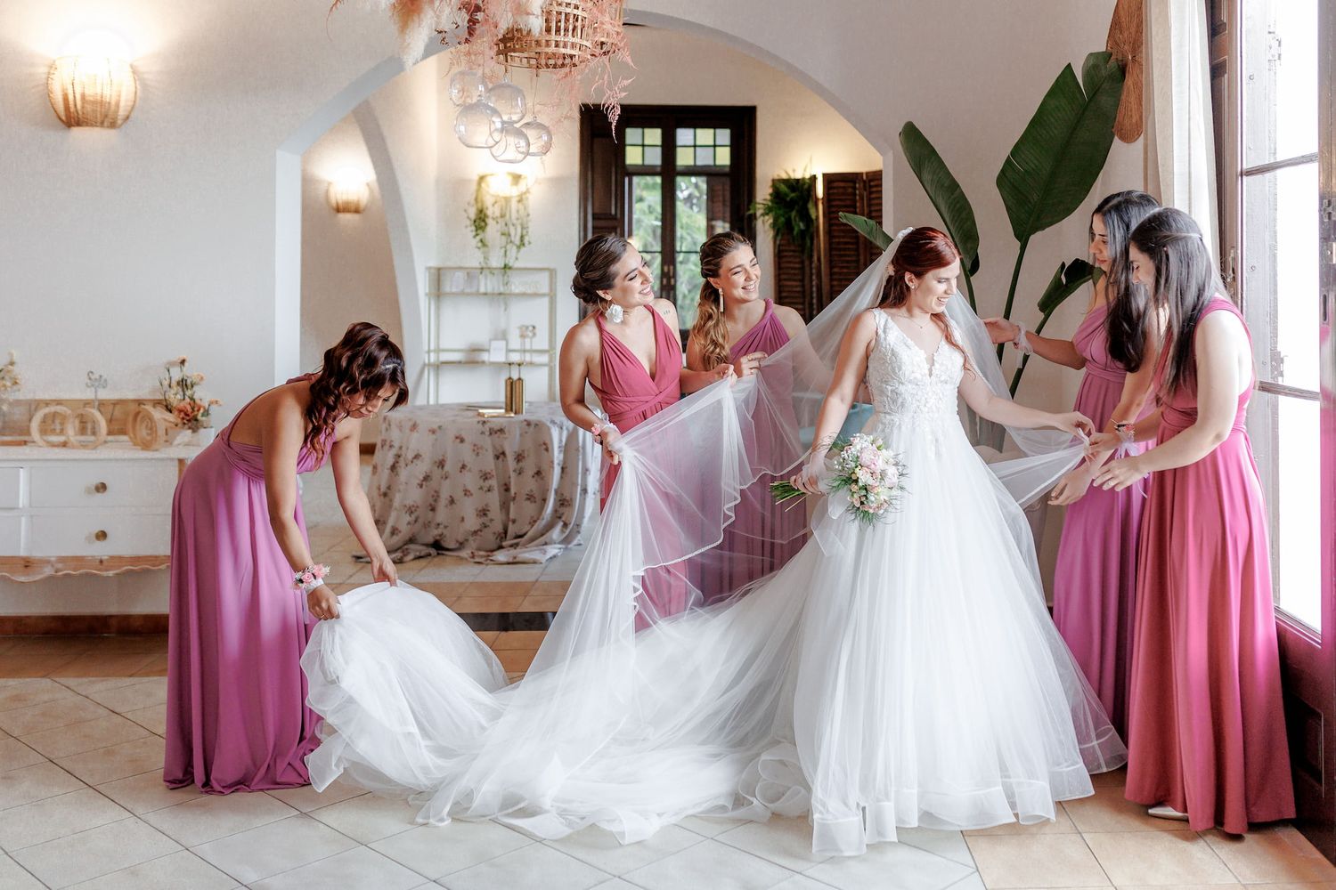 The bride is surrounded by her friends in pink dresses, and they adjust her veil and the designed wedding dress.