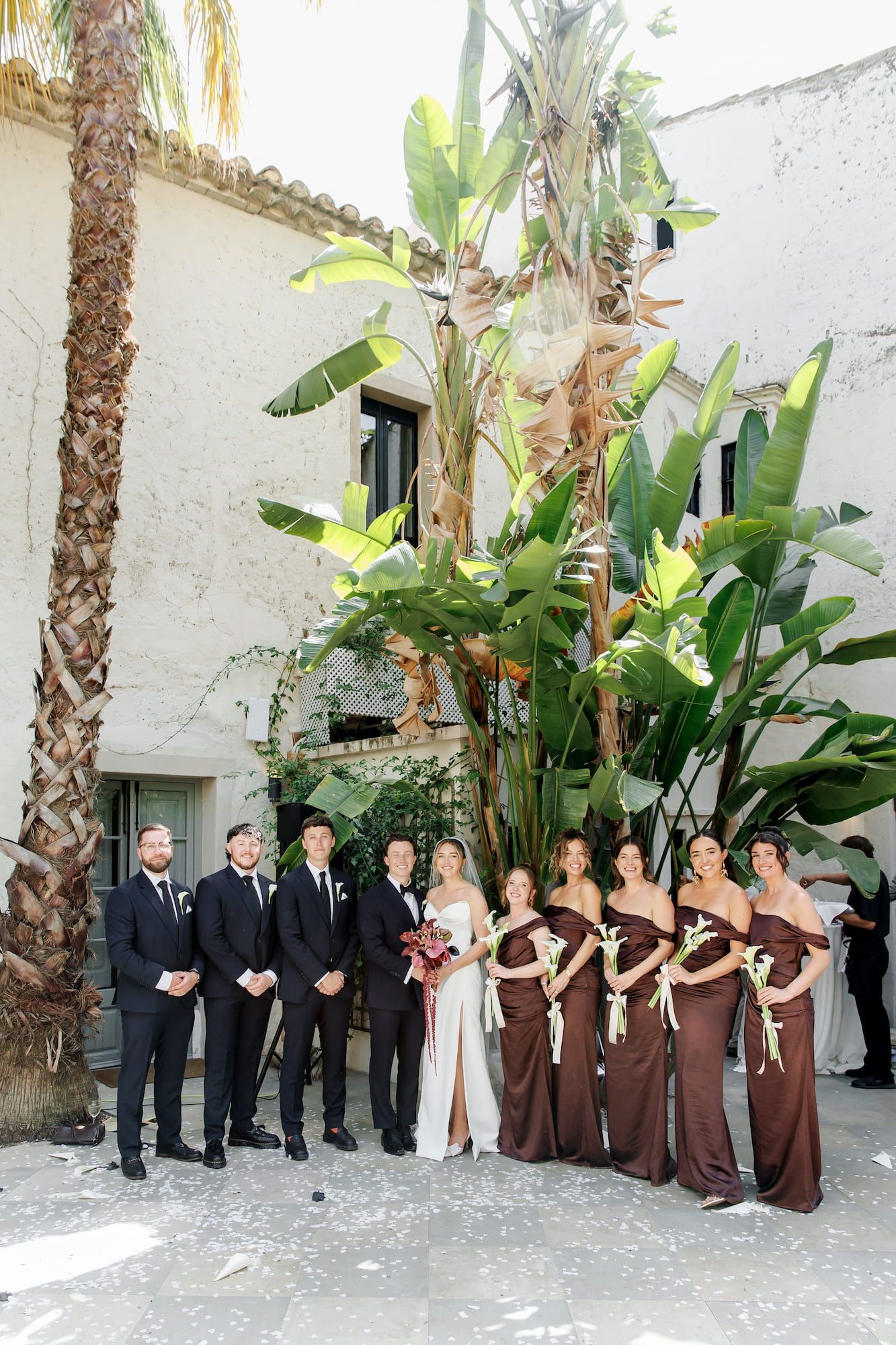 Wedding party poses outdoors with bridesmaids in brown gowns and groomsmen in black suits before a white building.