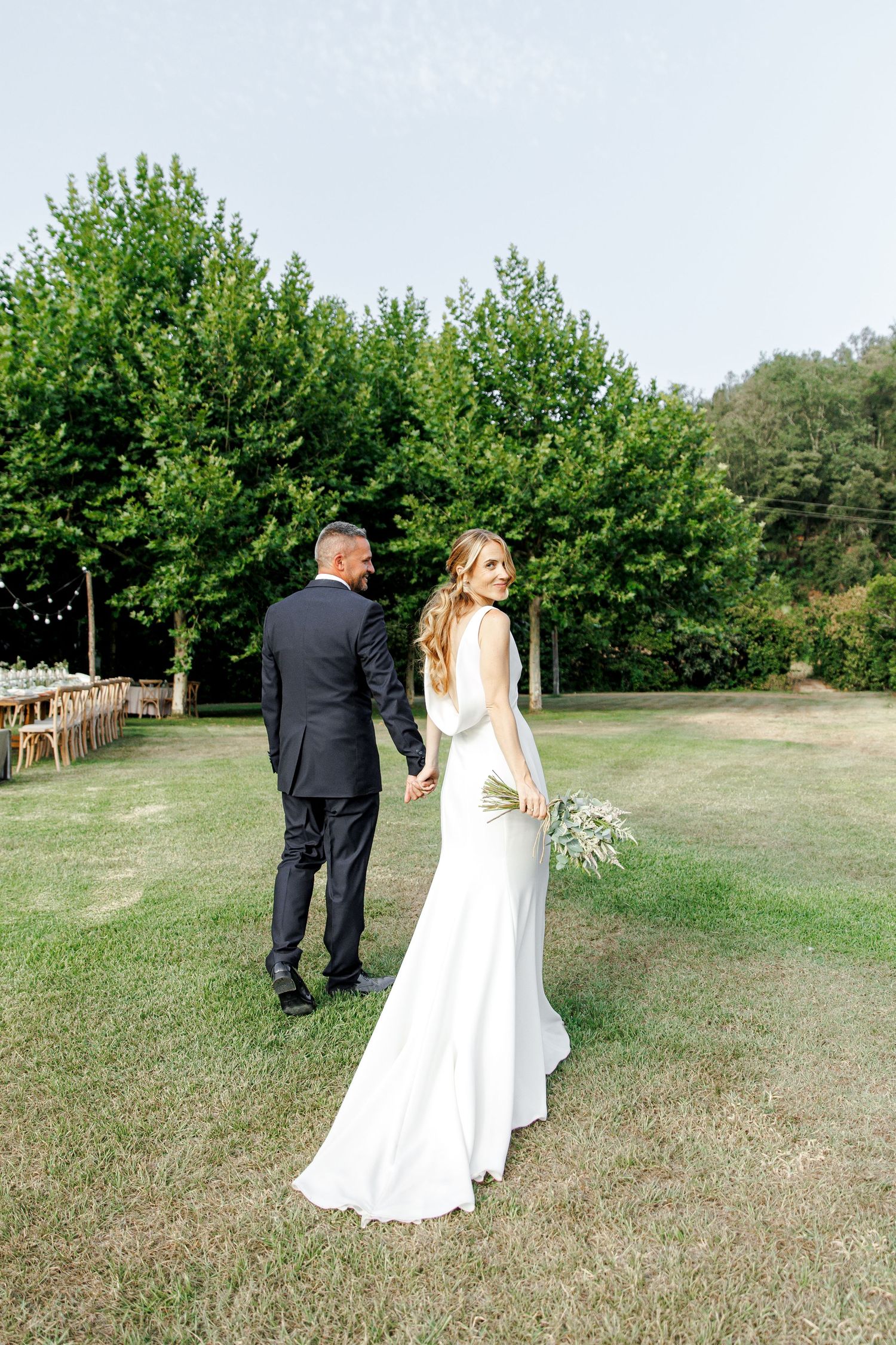 Bride in white gown looks back while holding groom's hand on green lawn with trees and reception tables in background.