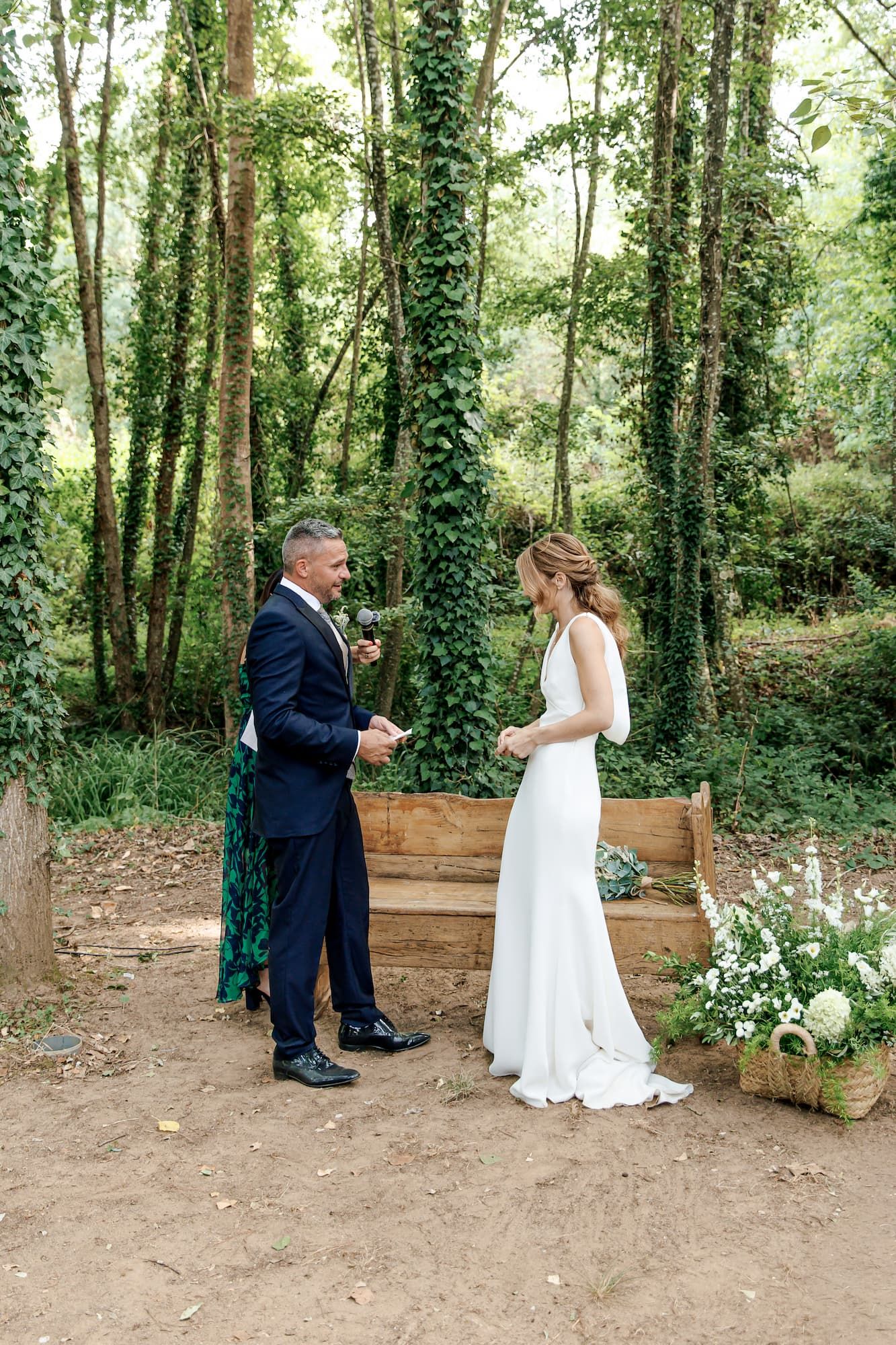 Bride and groom exchange vows outdoors in a lush forest garden setting, she in white gown, he in navy suit.