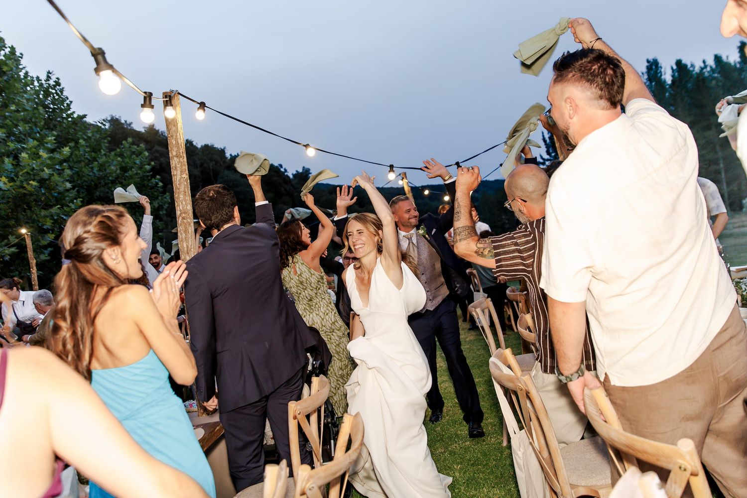 Bride and groom dancing joyfully with guests at outdoor wedding reception under string lights at dusk.