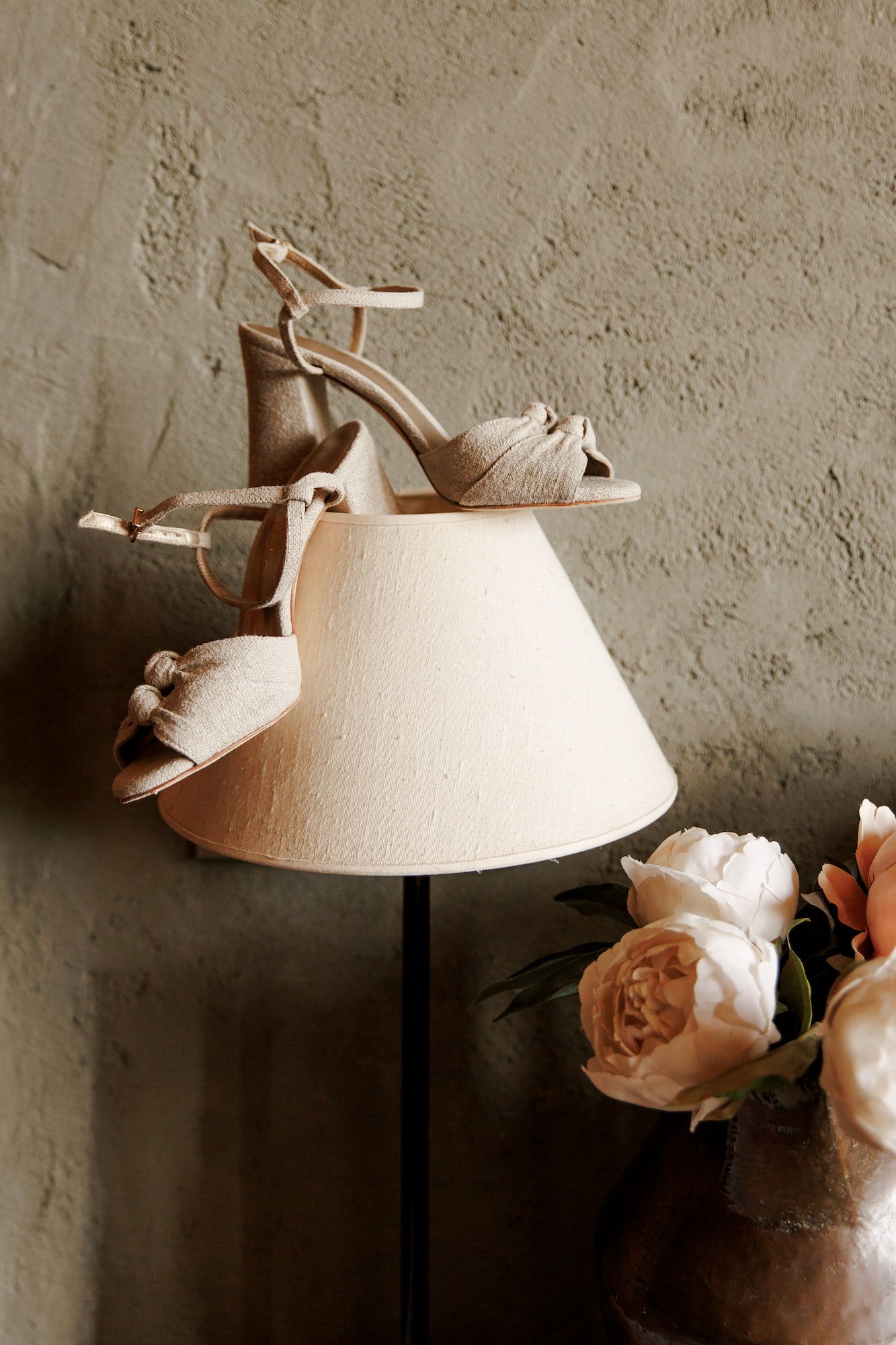 Elegant beige bridal heels with ankle straps displayed on a lamp shade, surrounded by soft pink roses against a textured wall.