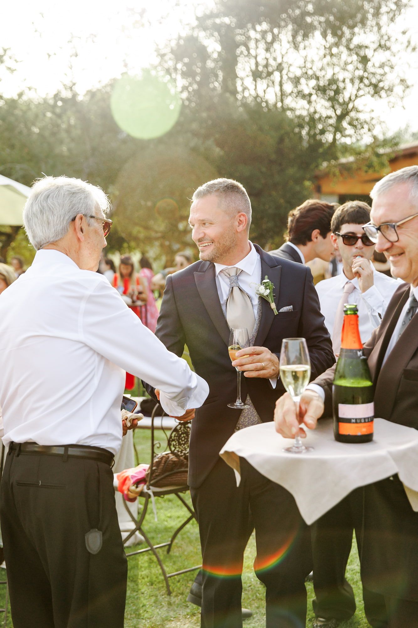 Groom in navy suit holding champagne glass at outdoor wedding reception with guests gathered around a table.