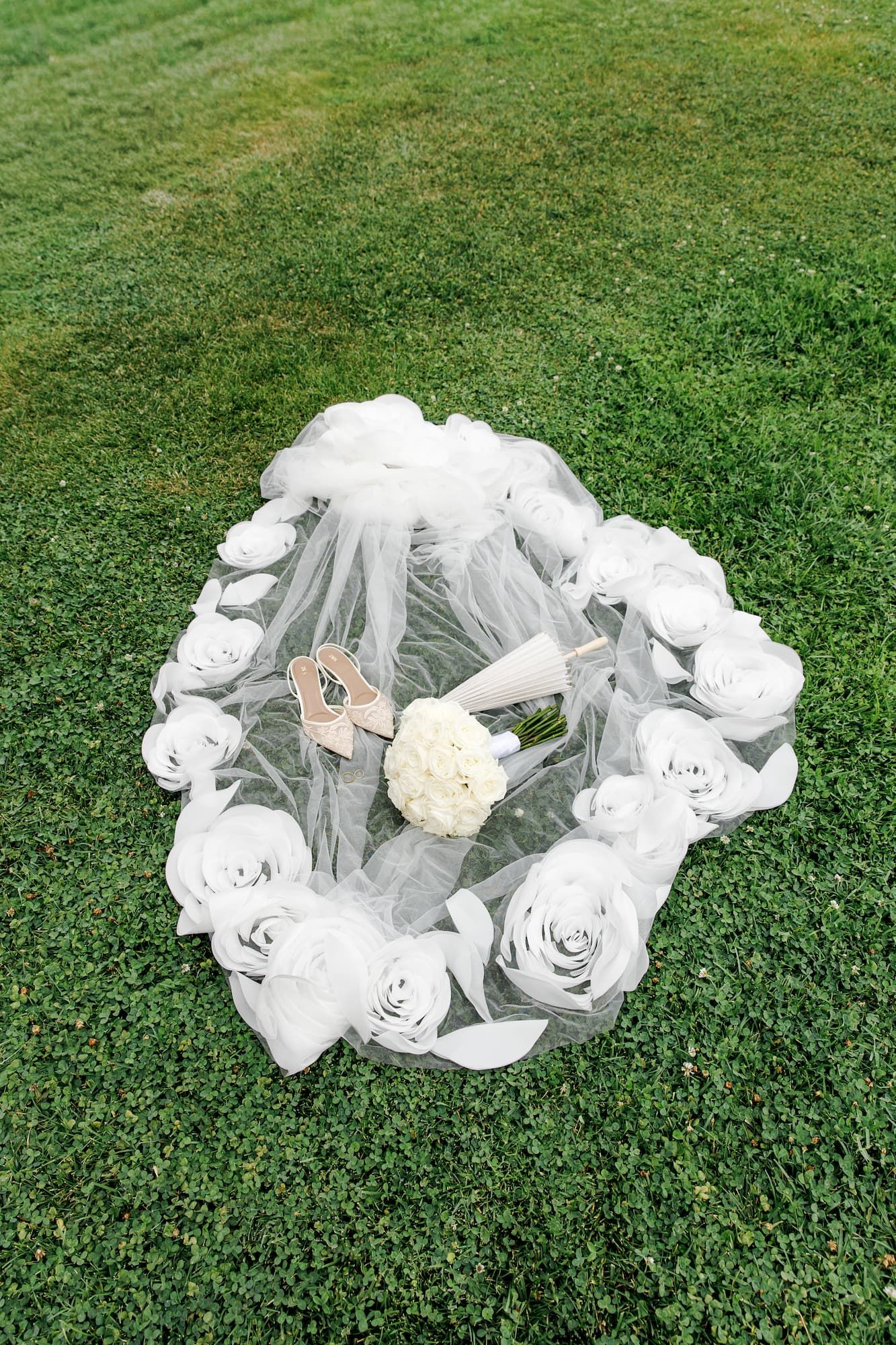 White bridal veil with paper roses and bouquet arranged on green grass for wedding photography.