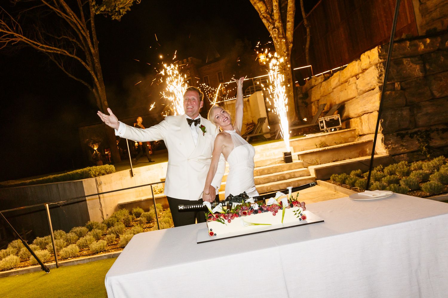 Bride and groom smiling beside a floral-topped wedding cake on a white-draped table at a nighttime outdoor reception with sparklers.