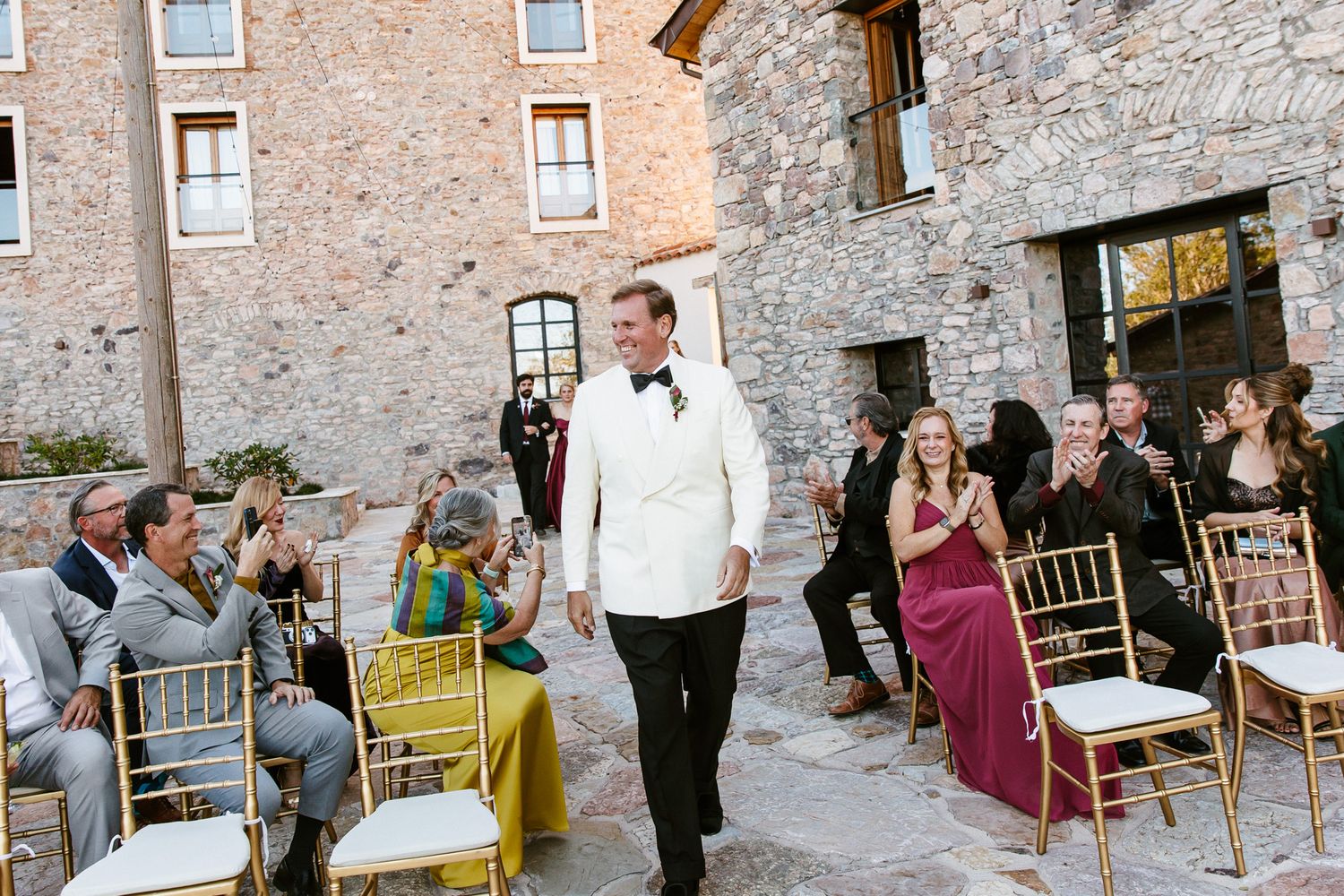 Groom in a white dinner jacket and bow tie walks down the aisle as seated wedding guests smile in a rustic stone courtyard.
