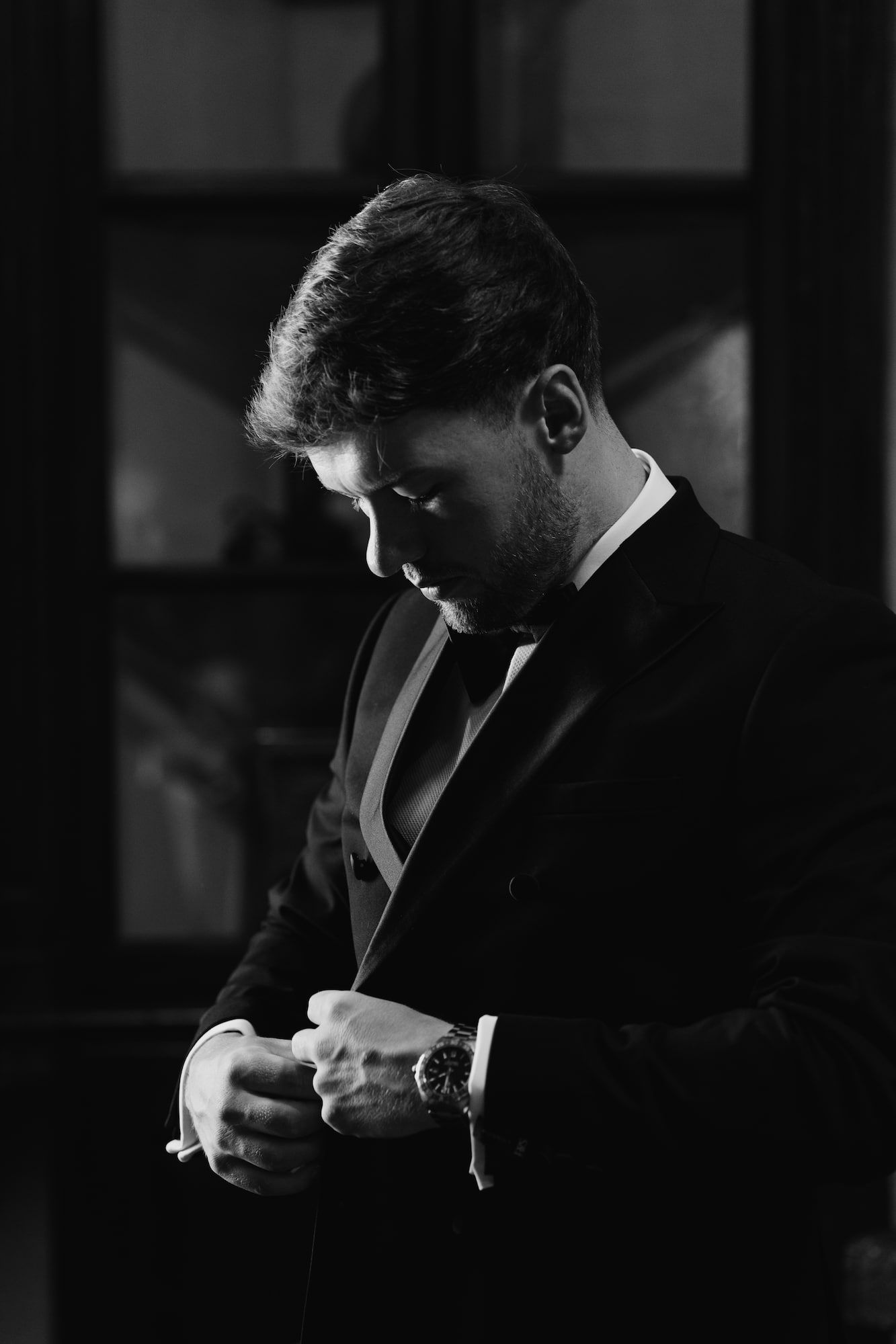 Elegant man in tuxedo and bow tie adjusting cufflinks, black and white portrait photography.