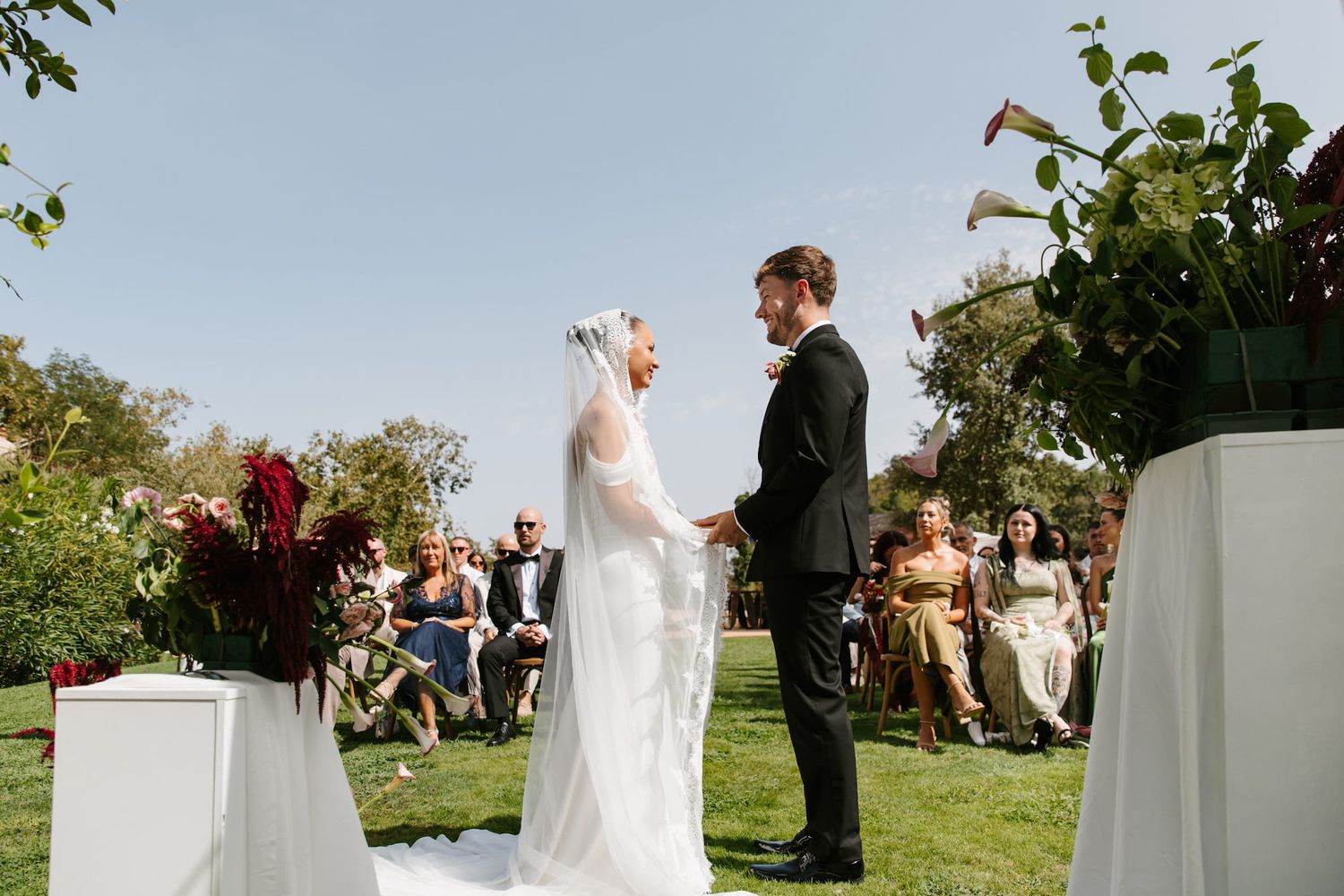 Bride and groom hold hands during outdoor wedding ceremony on sunny day with guests seated in background.