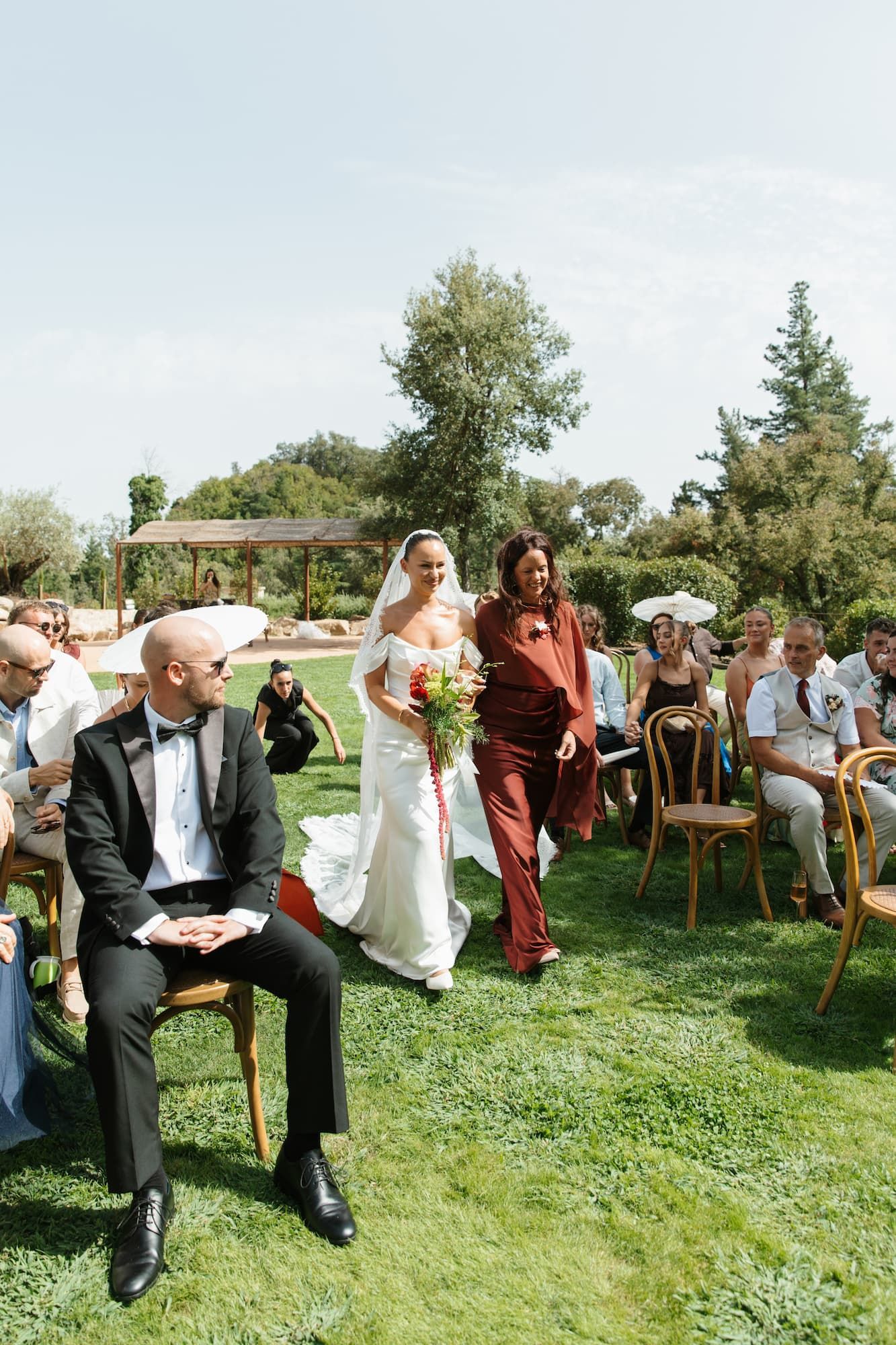 Bride in white gown walks down outdoor aisle with escort, groom seated in tuxedo, sunny garden wedding ceremony.