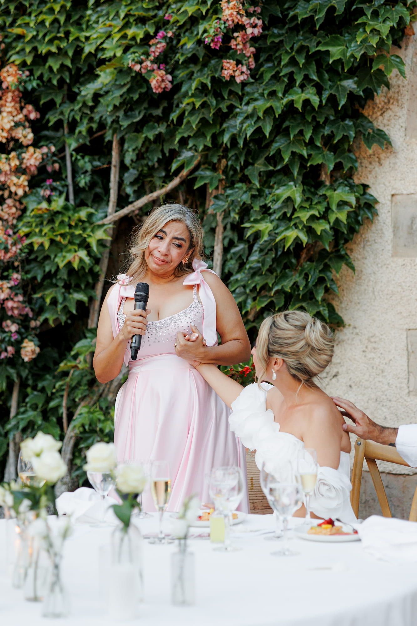 Bridesmaid in pink dress gives emotional speech with microphone at outdoor wedding reception with ivy backdrop.