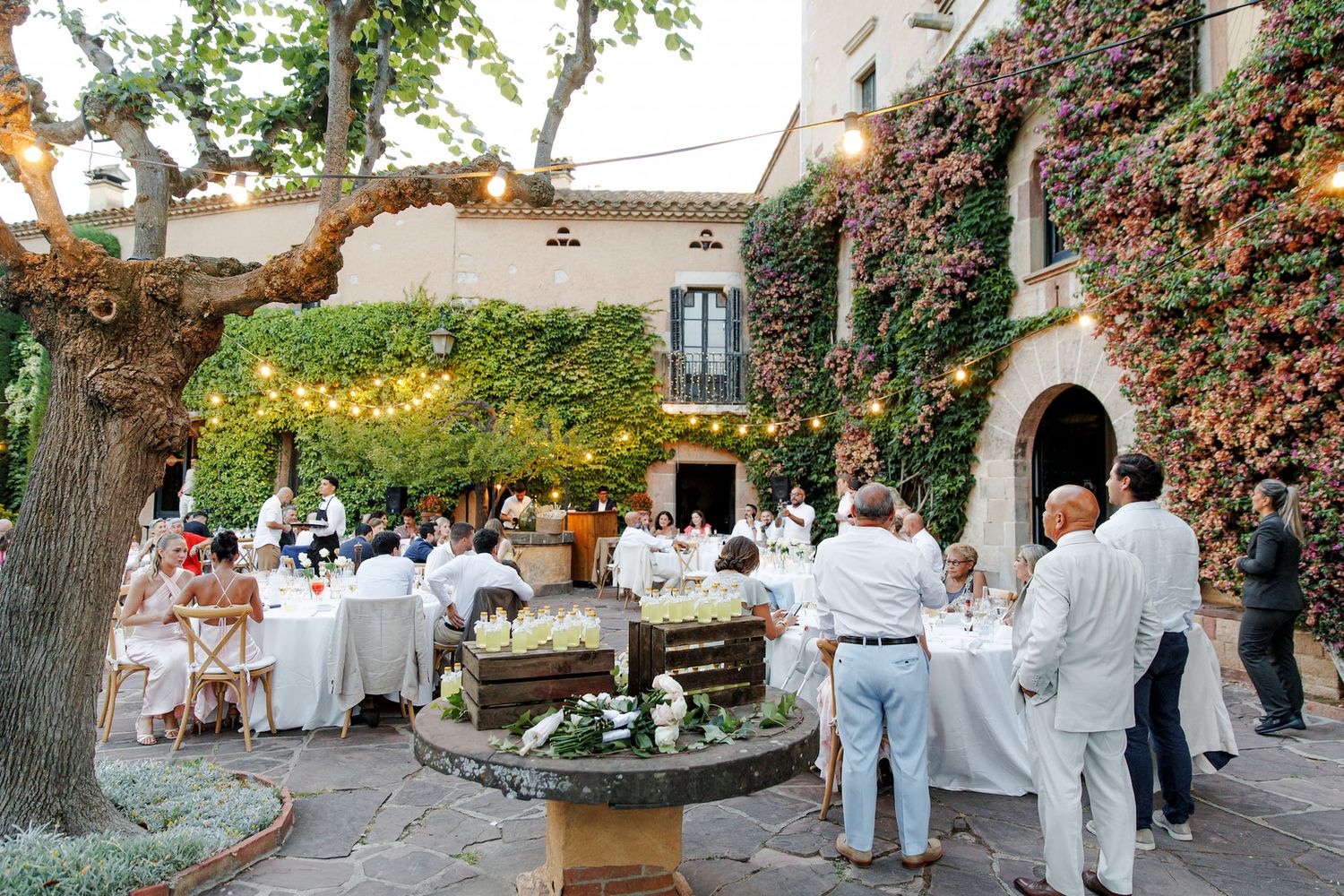 Elegant outdoor wedding reception in ivy-covered courtyard with string lights, white-draped tables, and guests in formal attire.