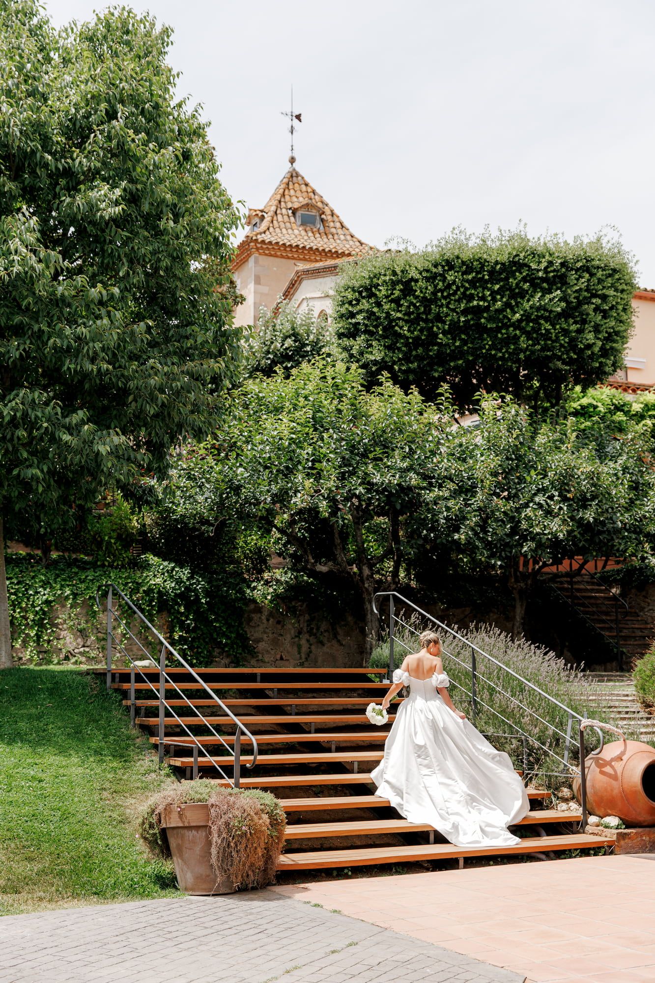 Bride in white gown ascending wooden stairs in lush garden with historic building backdrop.