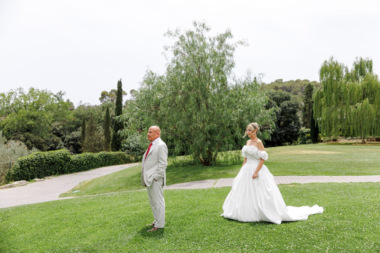 Bride in white gown and groom in gray suit share a first look moment on lush green lawn surrounded by trees.