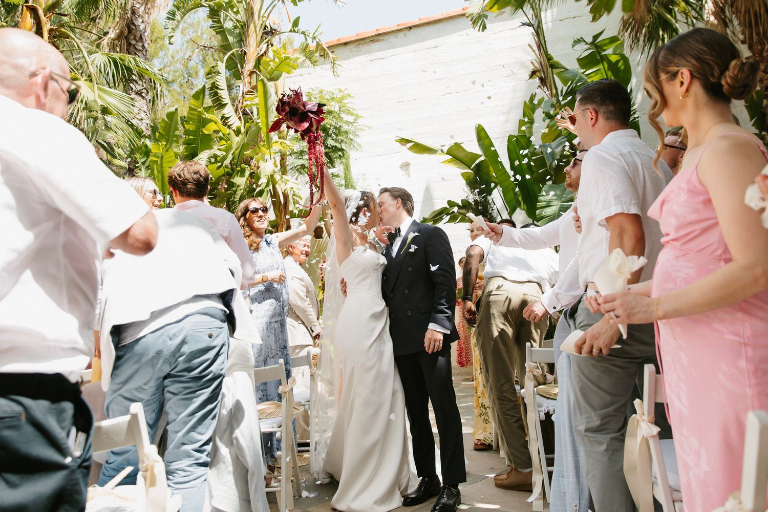 Bride and groom kiss while guests toss flower petals during outdoor wedding ceremony at Masia Victoria near Barcelona