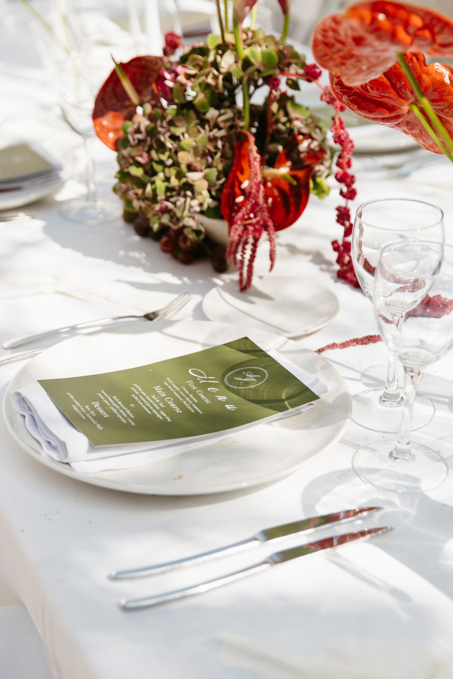 Elegant wedding table setting with white linen, green menu card, and red floral centerpiece.