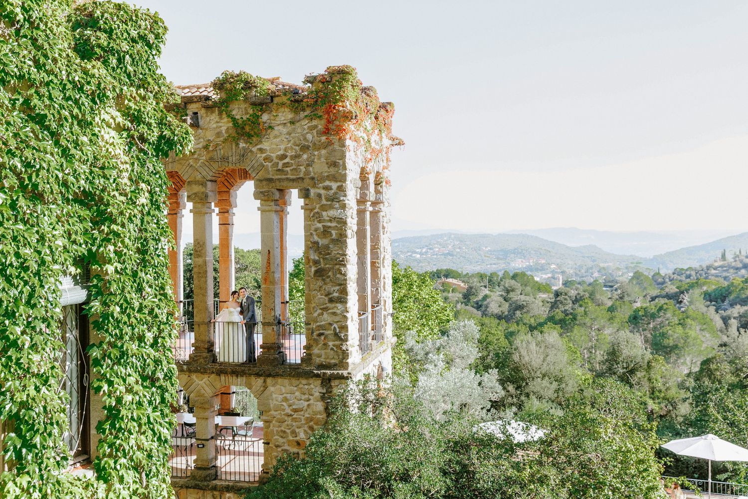 Ivy-covered La Baronia balcony with columns overlook a lush green valley and rolling hills under a clear sky