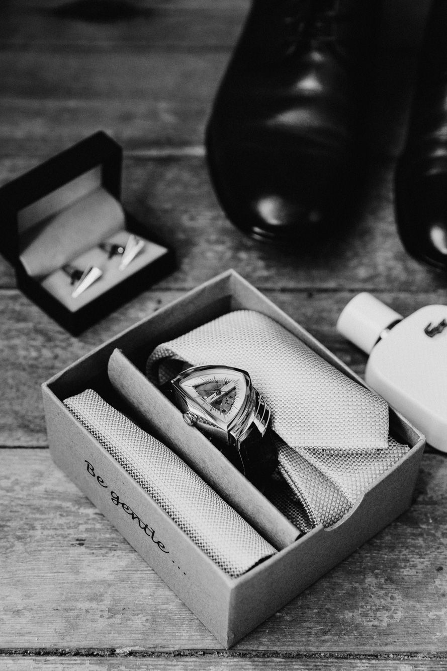 Black and white photo of a gift box with a watch, tie, cufflinks, cologne, and dress shoes for a groom.