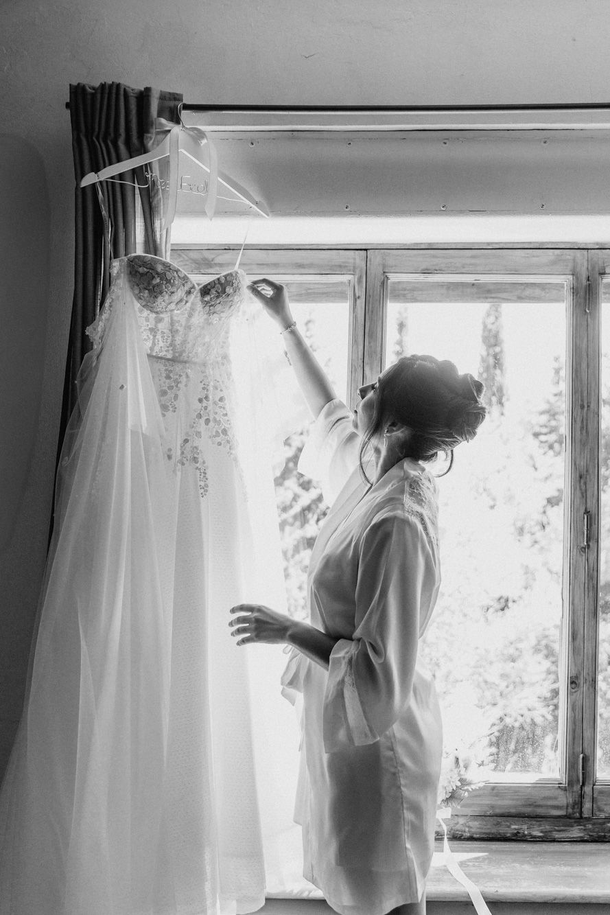 Black and white photo of a bride in a robe admiring her wedding dress hanging by a sunlit window.