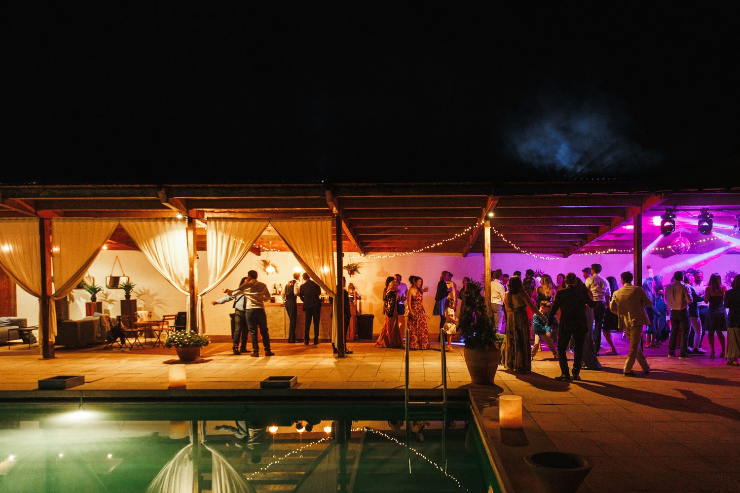 Outdoor nighttime pool party with guests dancing under warm string lights and vibrant purple DJ lighting.
