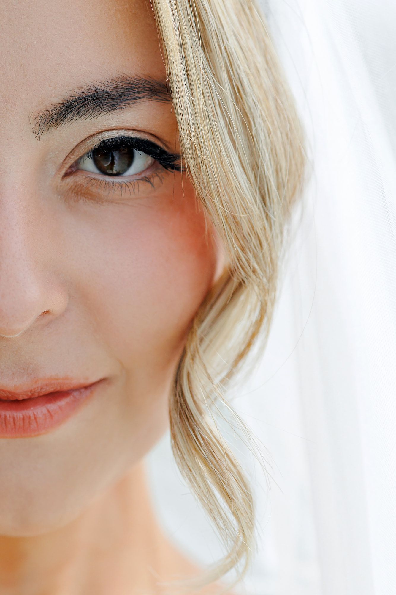 Close-up portrait of a blonde bride wearing a veil, showcasing elegant makeup with defined brows and eyeliner.