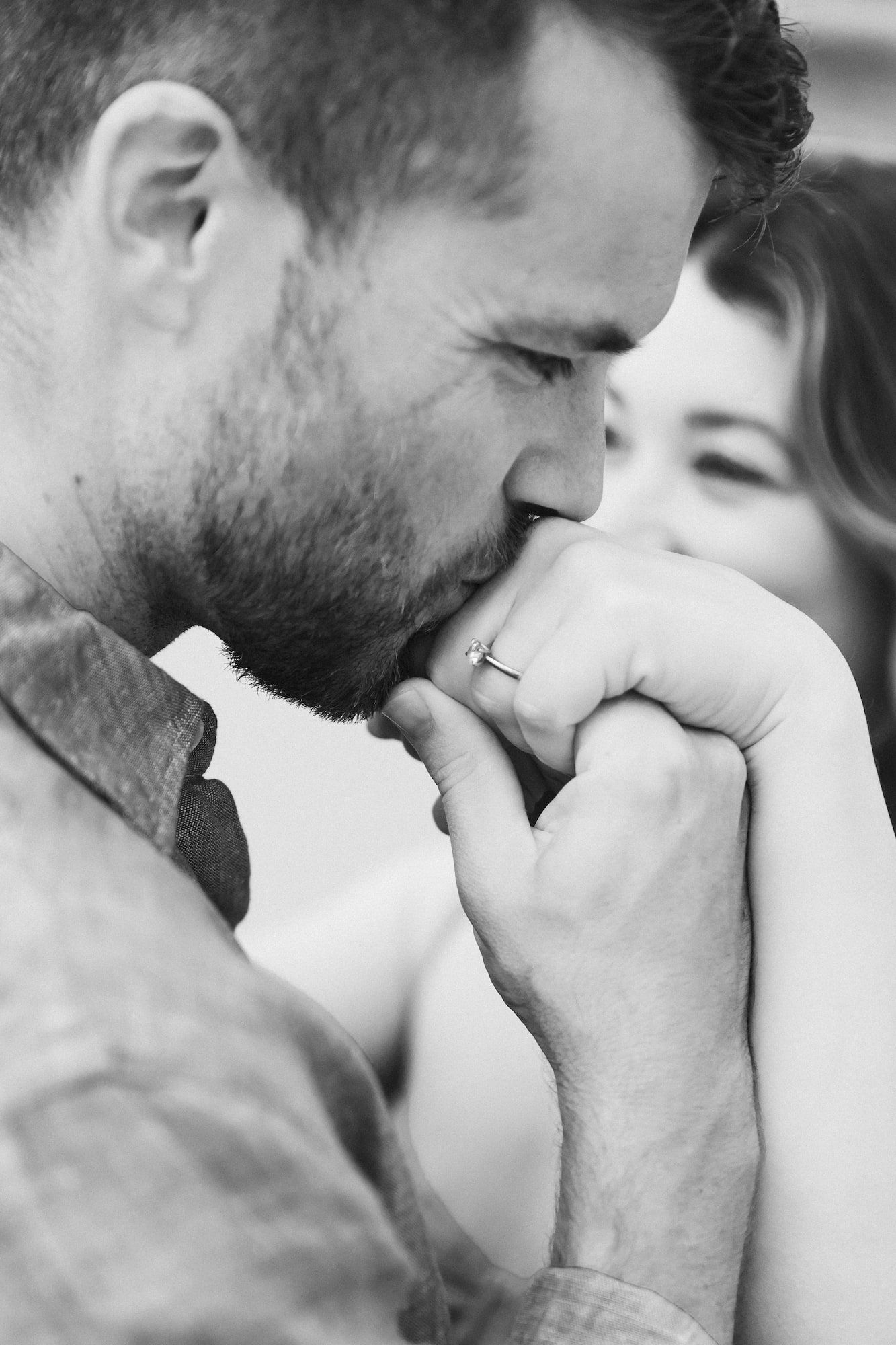 A man tenderly kisses a woman's hand as she smiles softly in this romantic black and white portrait.