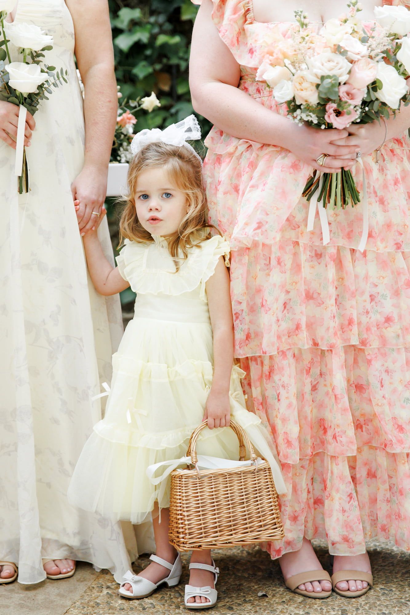 Flower girl in yellow ruffled dress holding wicker basket stands between bridesmaids in floral gowns at wedding.