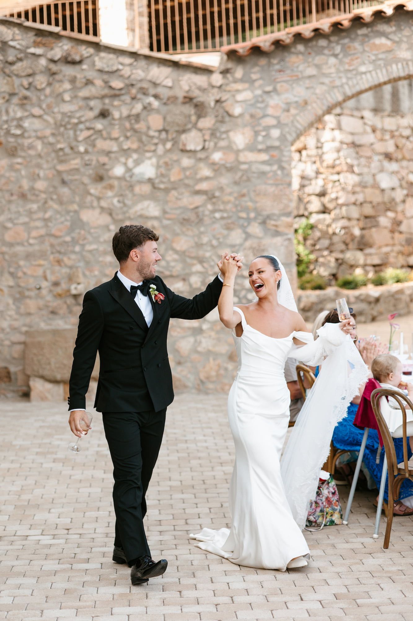 Bride and groom dancing joyfully outdoors at a rustic stone venue, bride in elegant white gown, groom in classic black tuxedo.