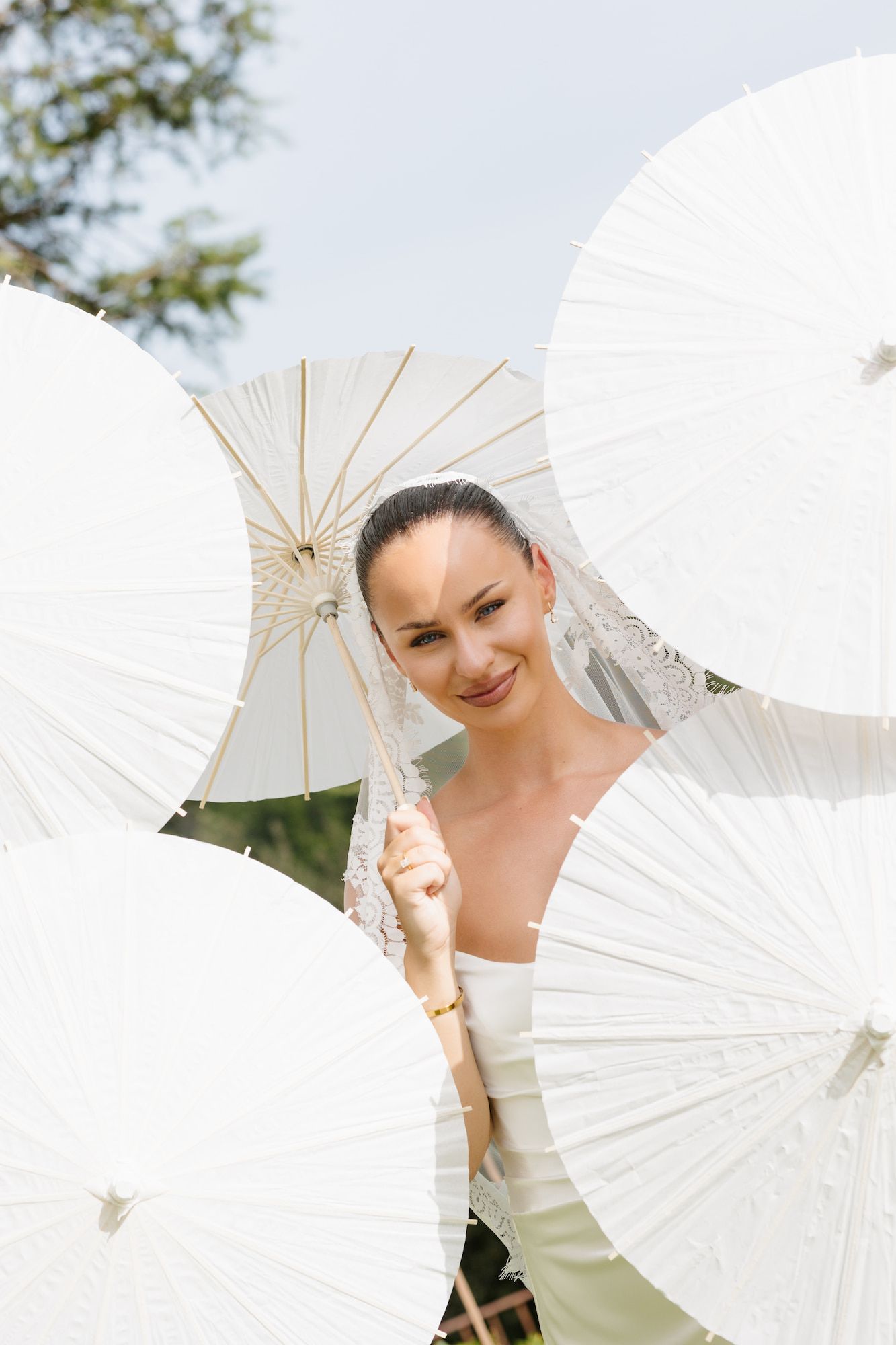 Smiling bride in white dress holding a paper parasol surrounded by multiple white umbrellas outdoors.