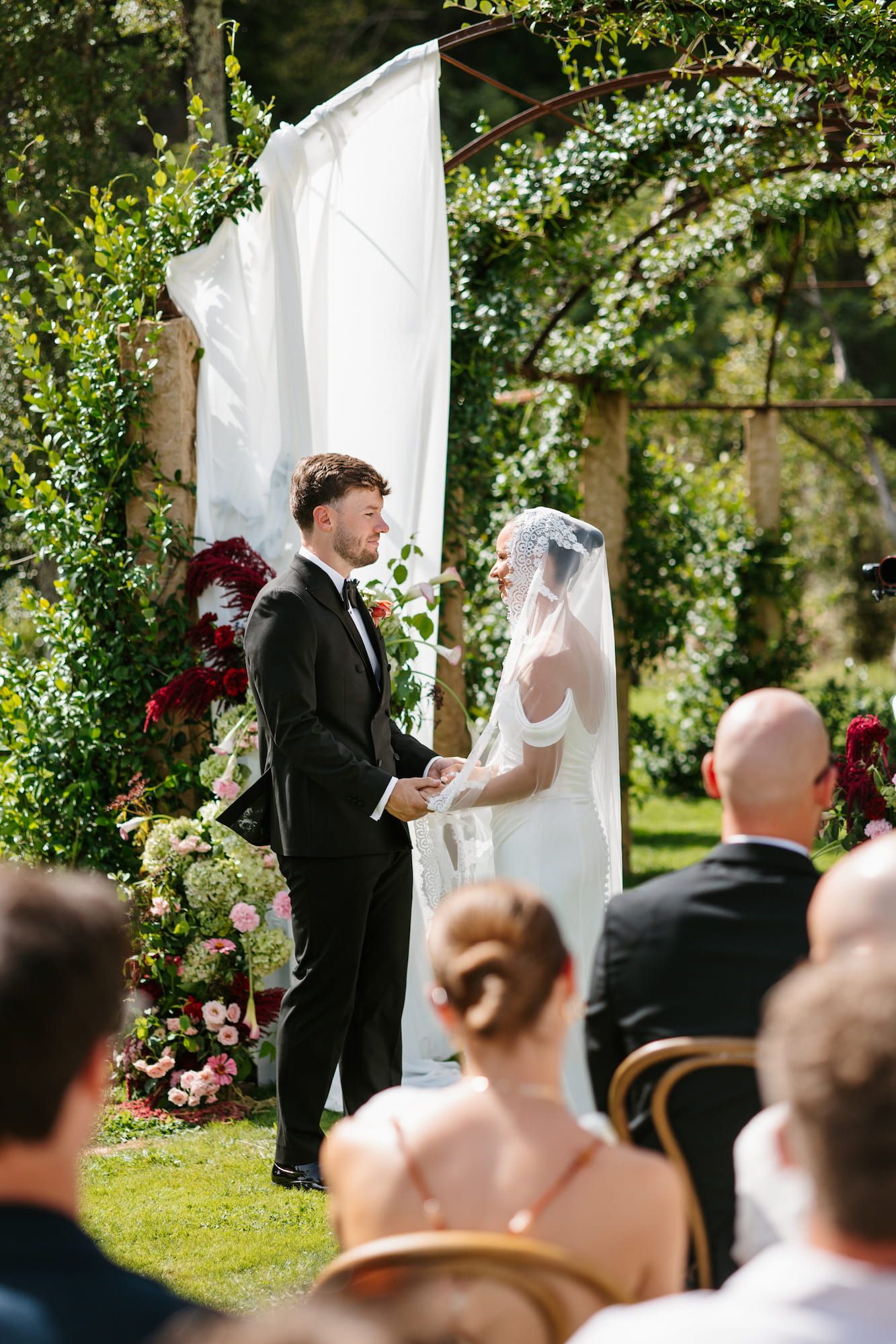 Outdoor wedding ceremony with groom in black tuxedo and bride in white gown exchanging vows under floral arch.