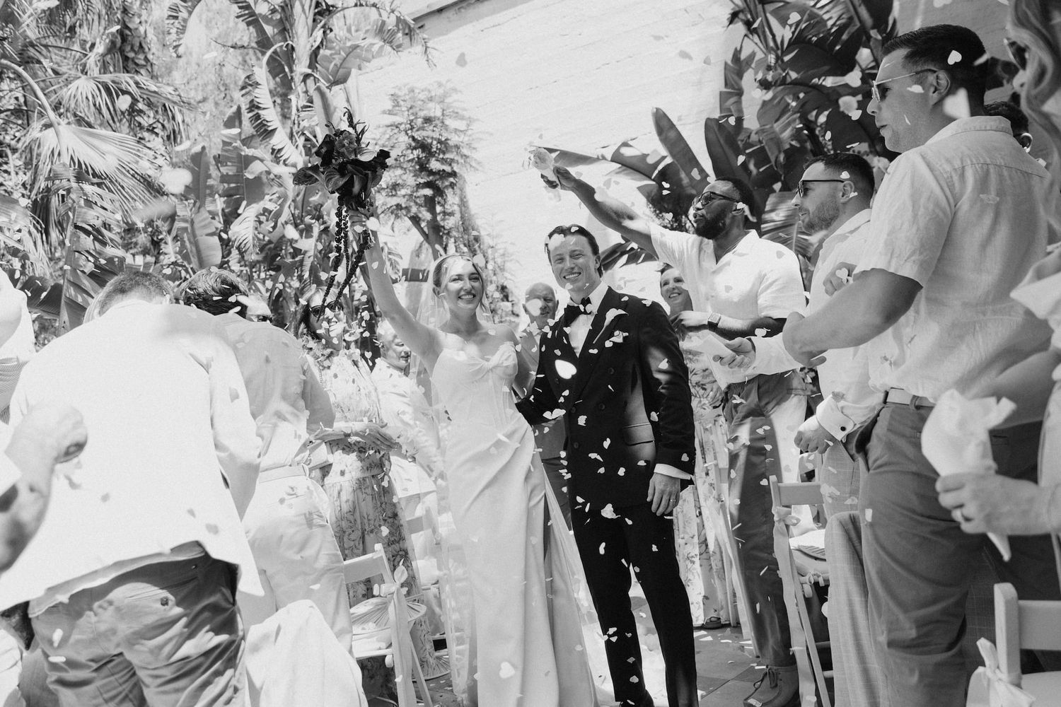 Black and white photo of a joyful couple walking down the aisle as guests toss confetti at their outdoor wedding.