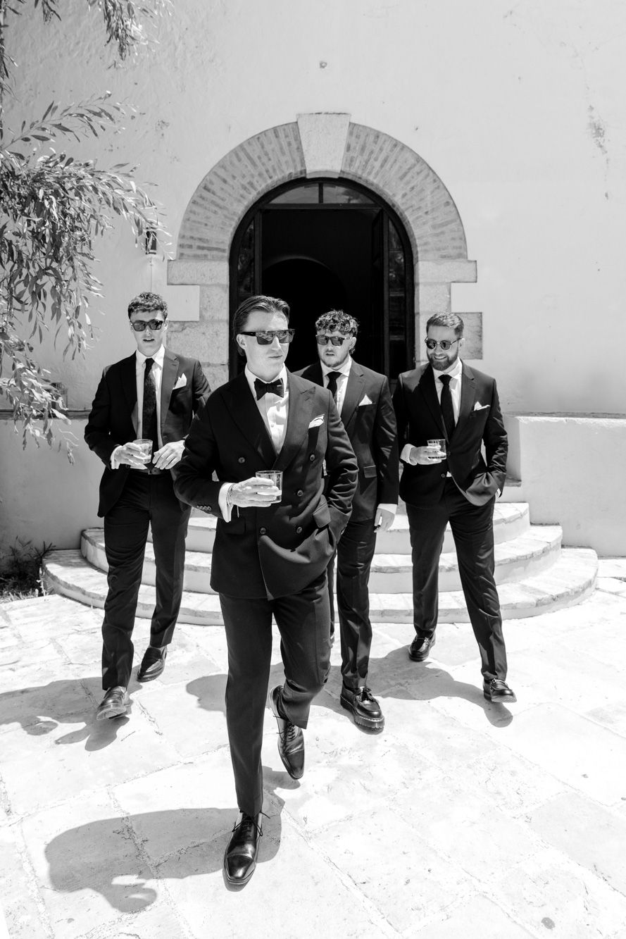 Four groomsmen in black suits and sunglasses pose confidently outside a stone church holding drinks.