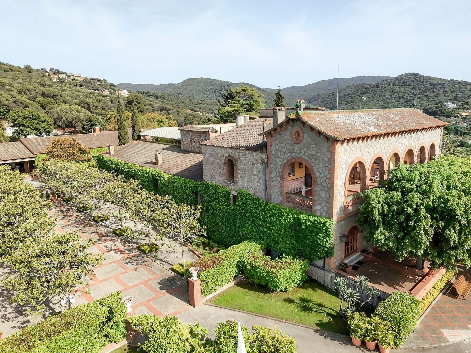 Aerial view of a stunning Mas de Sant Lleí wedding venue with arched windows, lush green hedges, and tree-covered hills in the background.